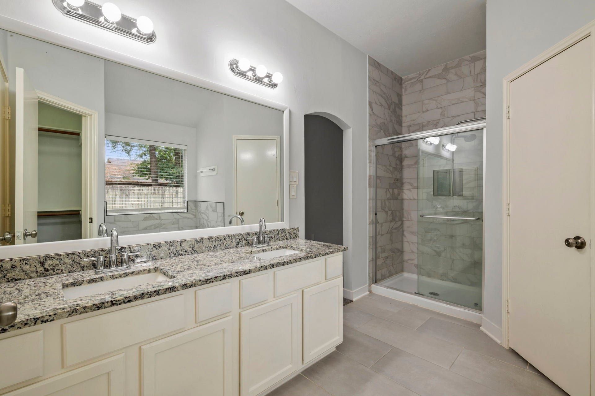 Elegant bathroom featuring a dual-sink granite countertop, stylish lighting, and a spacious glass-enclosed shower with modern tilework.