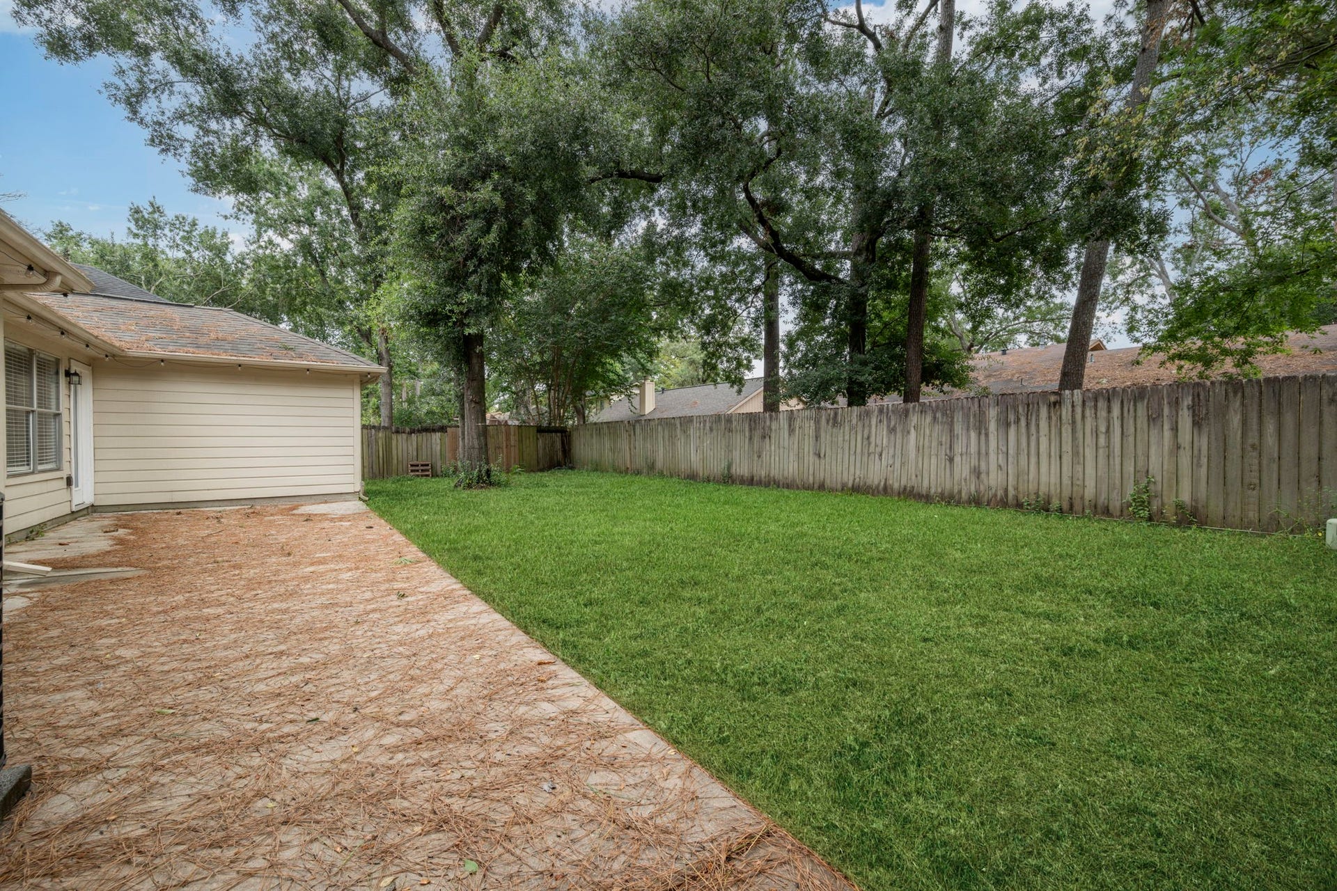 Spacious backyard featuring a lush green lawn, shaded by mature trees and complemented by a paved patio area perfect for outdoor entertaining.