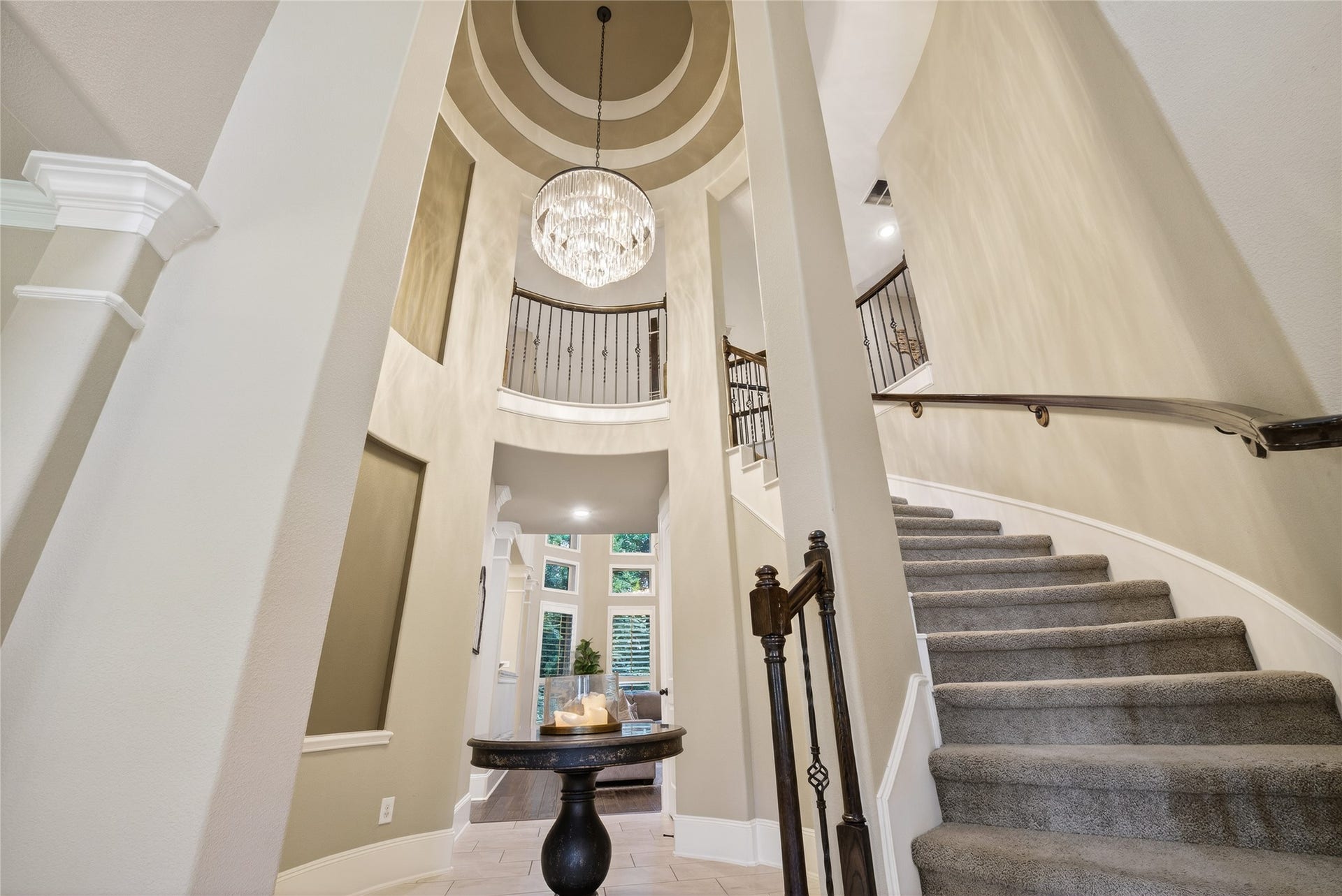 A grand chandelier hangs from a high ceiling, illuminating the entryway. The space is bright and inviting, with neutral tones and a modern aesthetic.