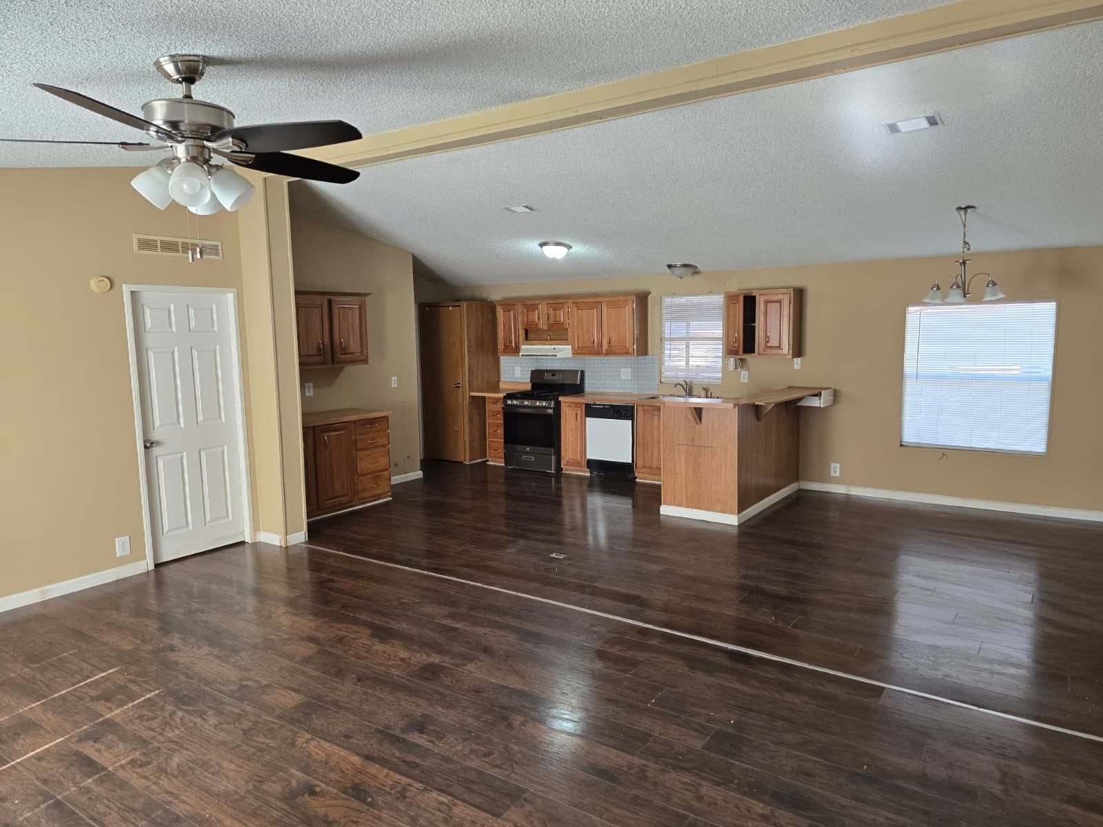 Spacious open-concept living area with rich wood flooring, a modern ceiling fan, and a cozy kitchen featuring ample cabinetry and natural light.