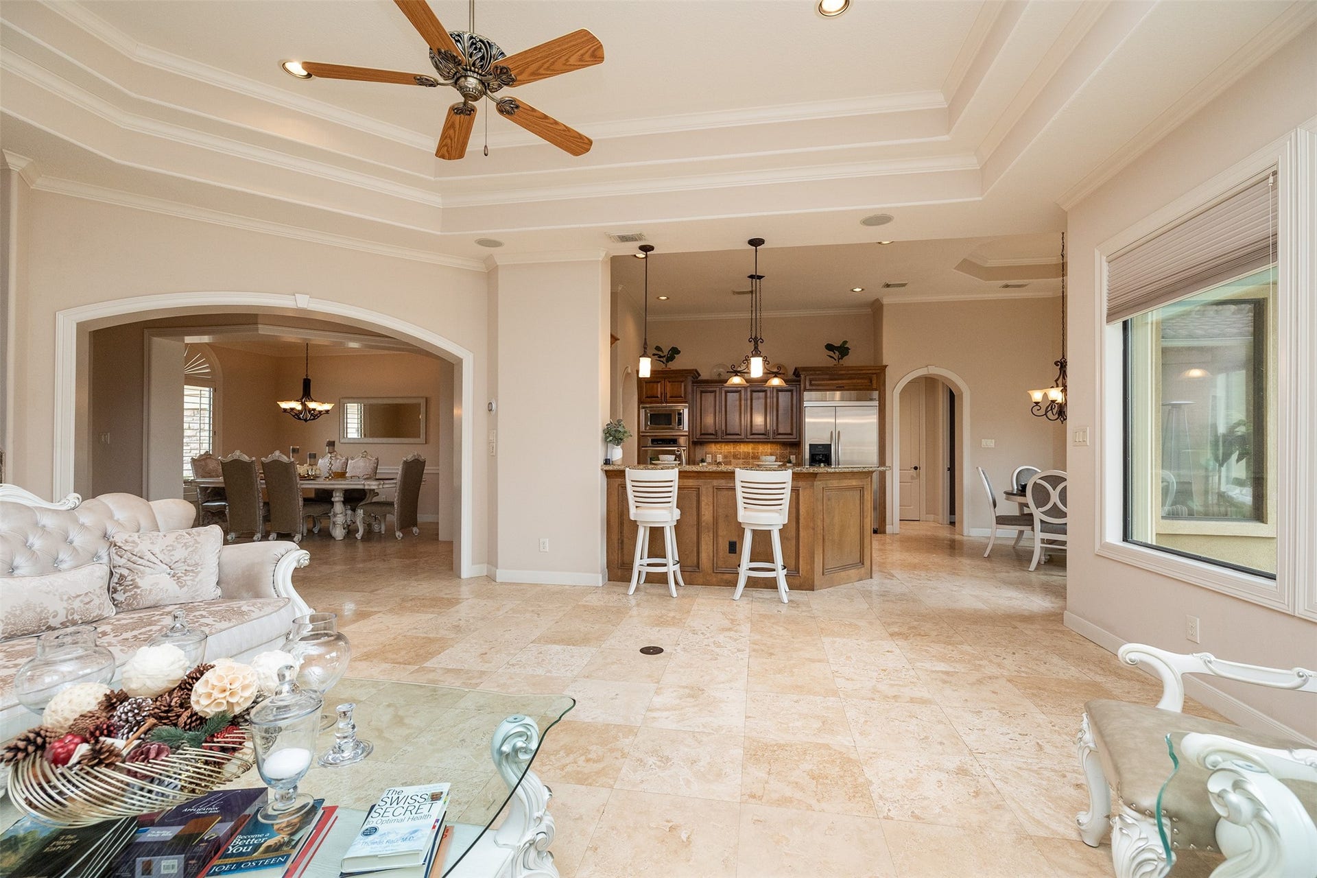 This elegant open-concept space features a bright living area with a coffered ceiling and ceiling fan, flowing into a modern kitchen with a large island and bar seating. Adjacent is a formal dining room, all with sleek tile flooring and ample natural light from large windows. Perfect for entertaining and family gatherings.