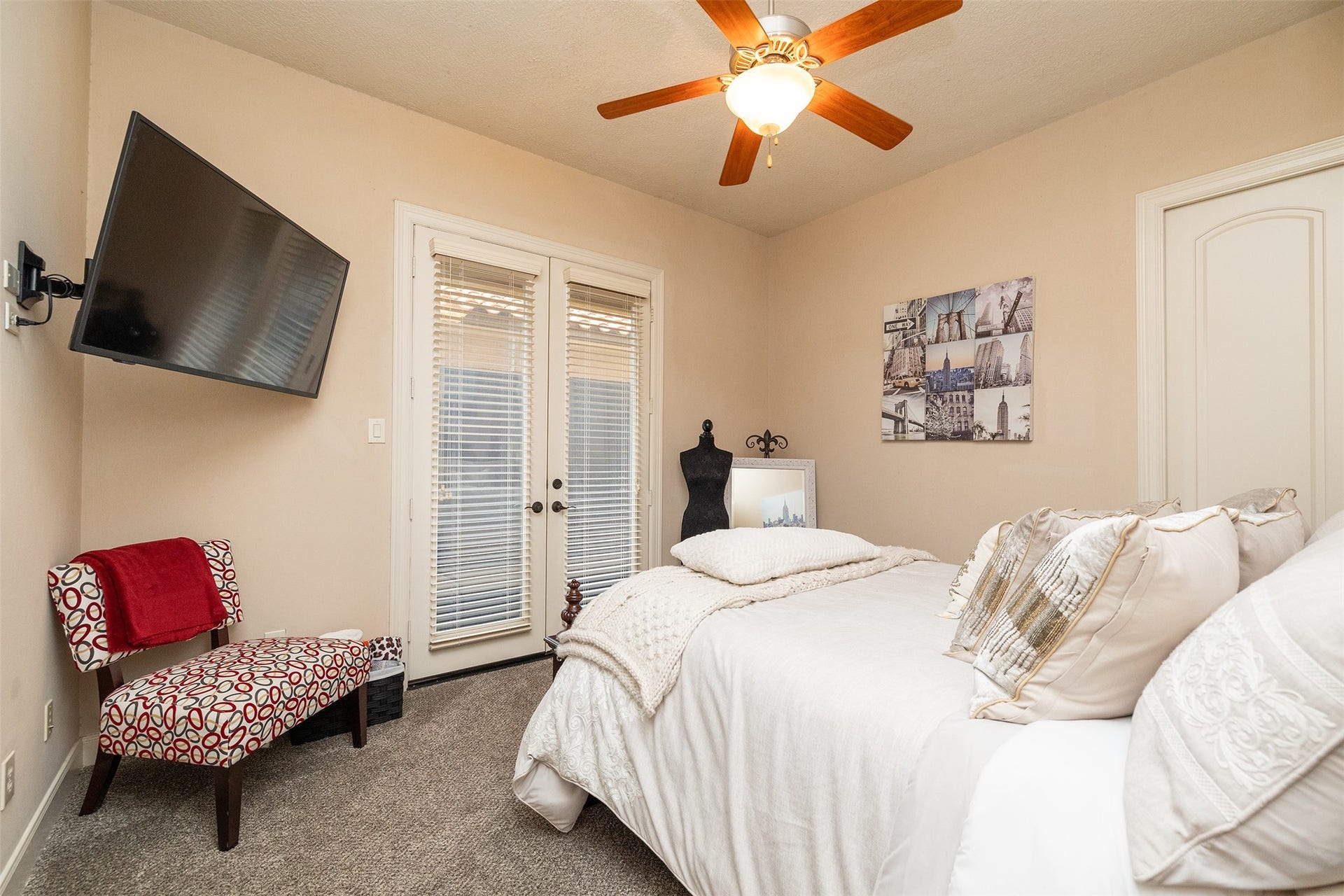 Cozy bedroom with a warm color palette featuring a ceiling fan, wall-mounted TV, and French doors that let in natural light. The room includes a stylish chair, decorative artwork, and plush bedding for a comfortable and inviting atmosphere.