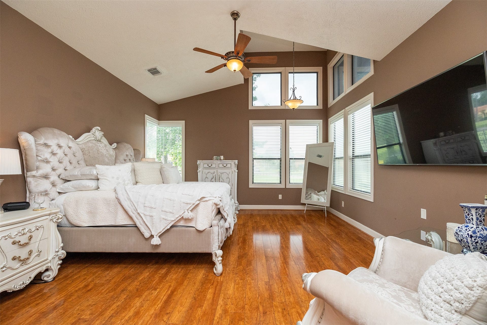 This spacious bedroom features elegant decor with a plush, tufted bed, rich wood flooring, and high ceilings. Large windows provide ample natural light, complemented by a ceiling fan and a wall-mounted TV for added comfort and entertainment.