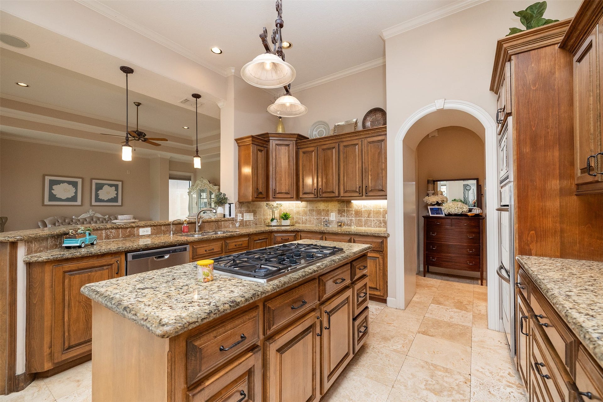 This spacious kitchen features elegant wood cabinetry, granite countertops, and a central island with a built-in gas cooktop. Recessed lighting and pendant fixtures provide ample illumination, while an archway leads to additional living space. The open layout connects seamlessly to the adjacent dining area, creating an inviting atmosphere for cooking and entertaining.