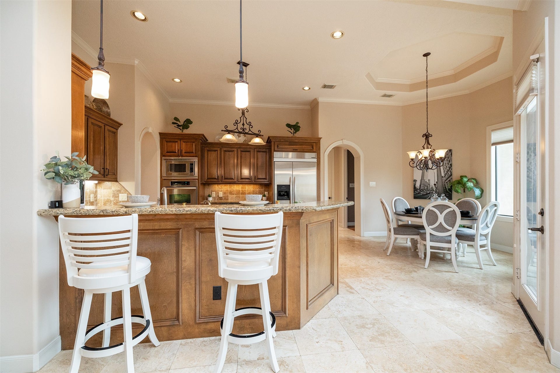 This photo showcases a spacious kitchen with a central island featuring granite countertops and stylish pendant lighting. The open layout includes a dining area with elegant chairs, neutral tones, and ample natural light from large windows.