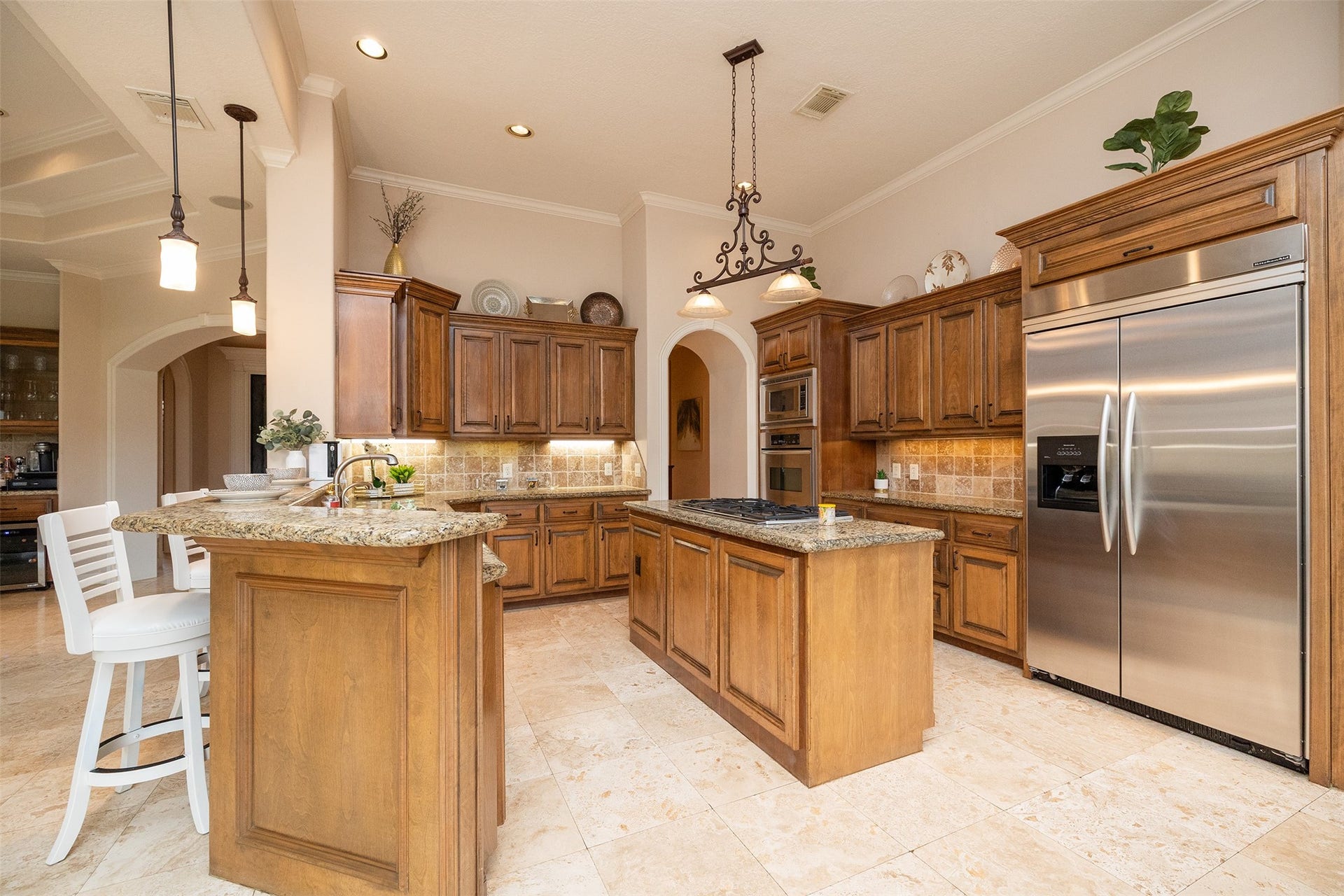 This photo showcases a spacious, elegant kitchen featuring warm wood cabinetry, granite countertops, and a large island with seating. Stainless steel appliances and pendant lighting add modern touches, while tile flooring completes the look.