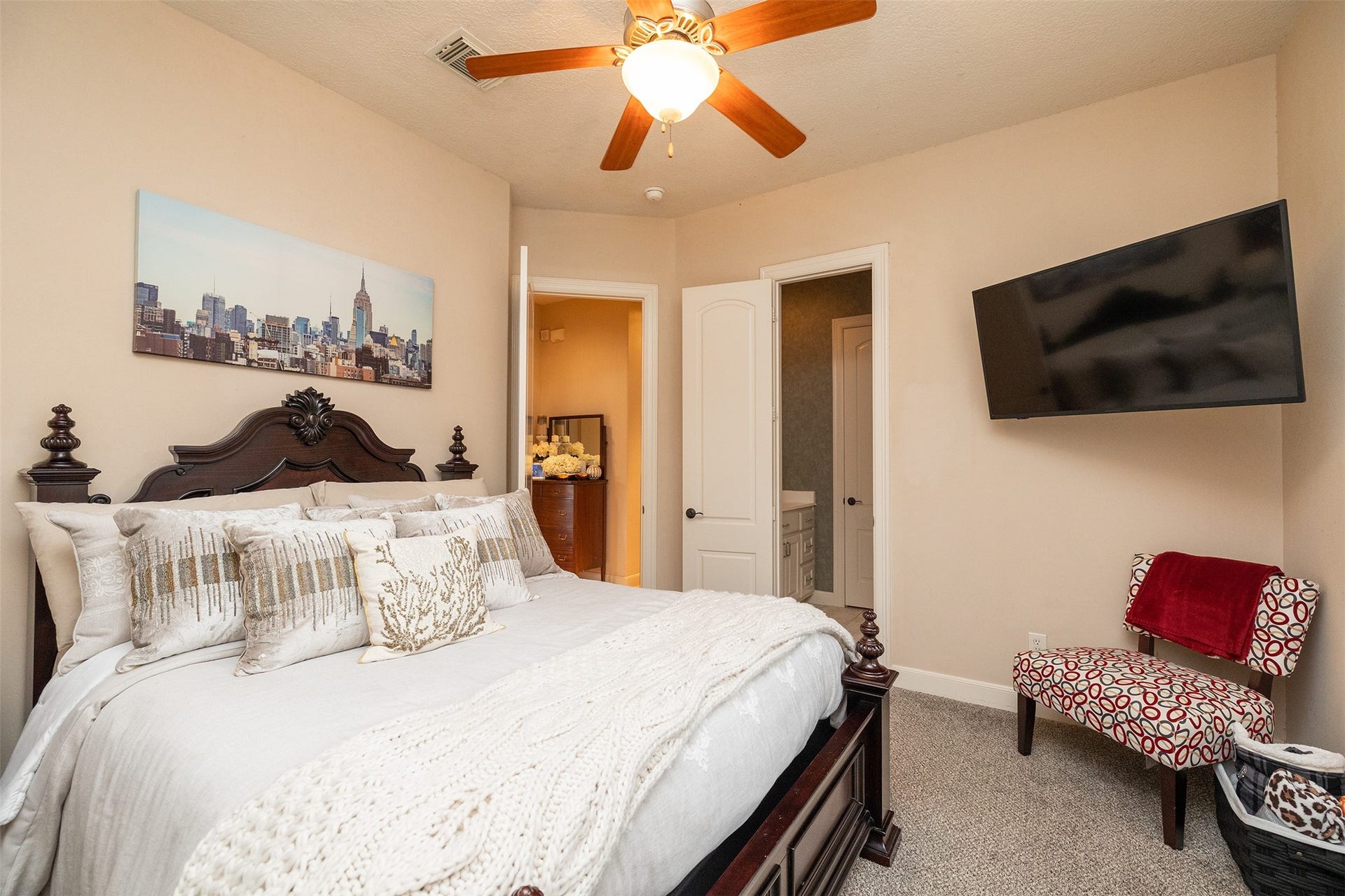 Cozy bedroom featuring a large bed with decorative pillows, a ceiling fan, wall-mounted TV, and a view into an ensuite bathroom. The space is warmly lit and tastefully decorated.