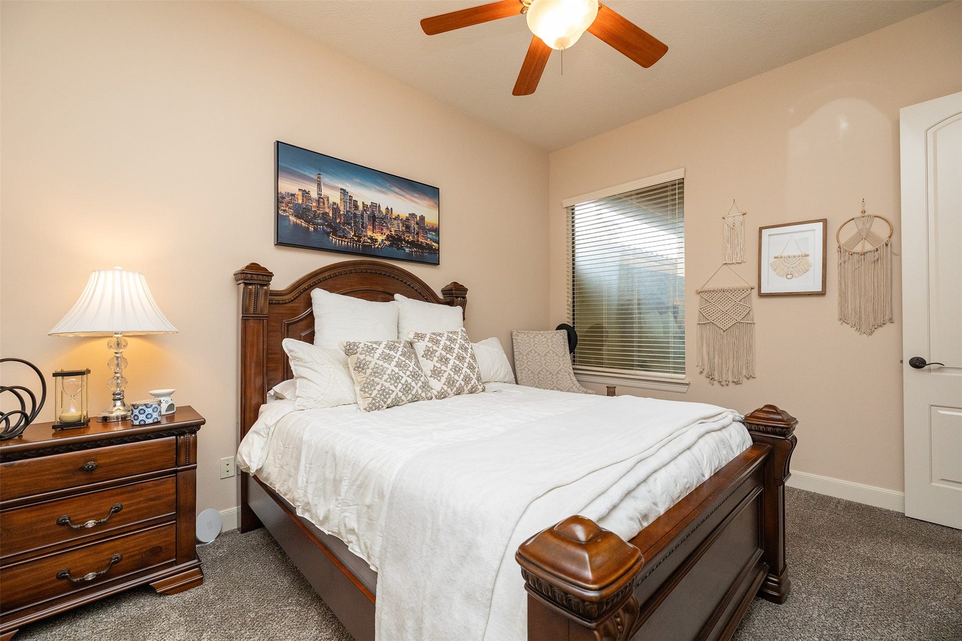 Cozy bedroom with warm neutral walls, featuring a wooden bed frame and matching nightstand. A cityscape artwork adorns the wall, complemented by soft lighting from a stylish lamp. A ceiling fan and window provide ventilation and natural light.