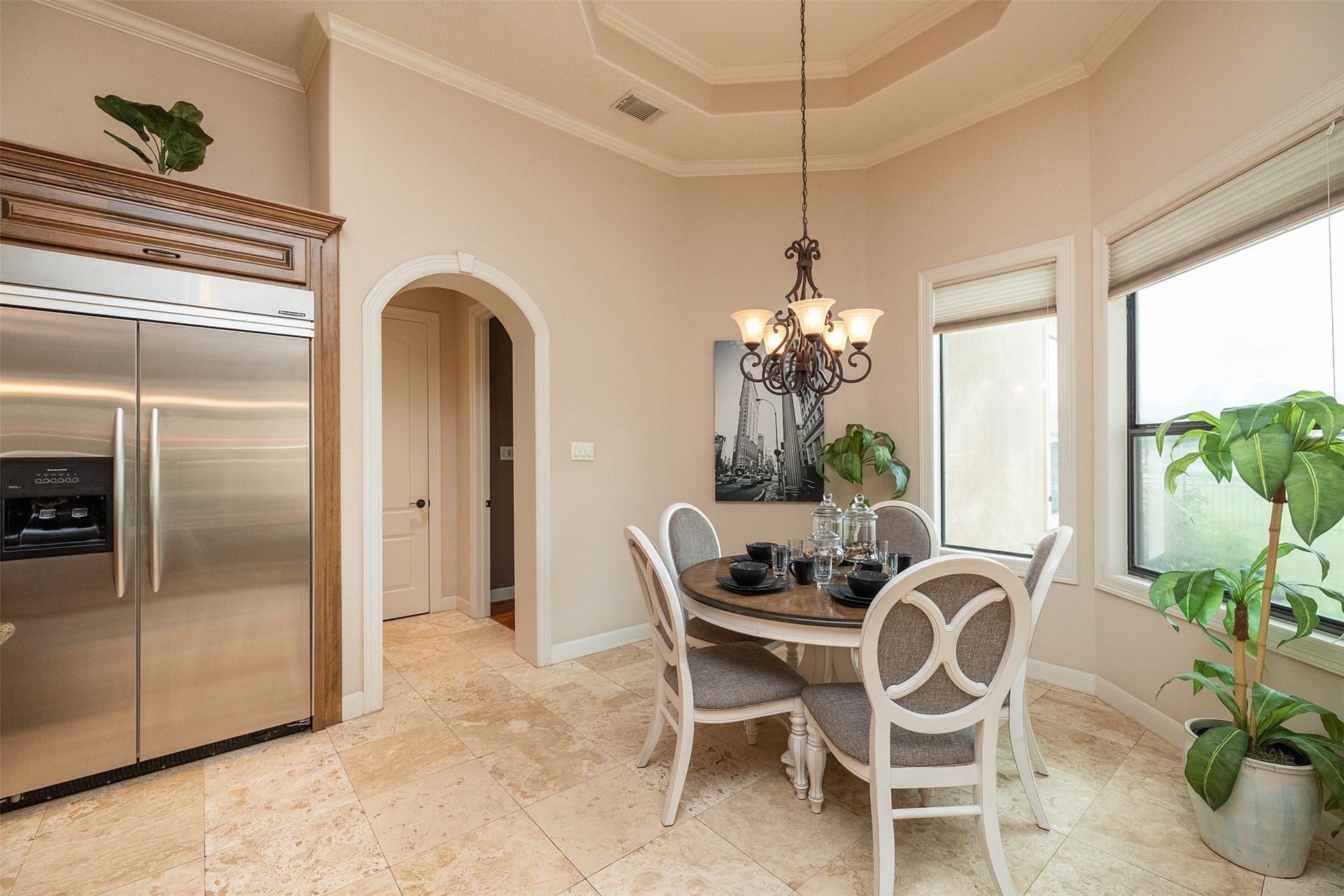 Bright dining area with elegant chandelier, large windows, and a cozy round table. Features a stainless steel fridge and archway leading to another room. Neutral tones and tiled floor add a modern touch.
