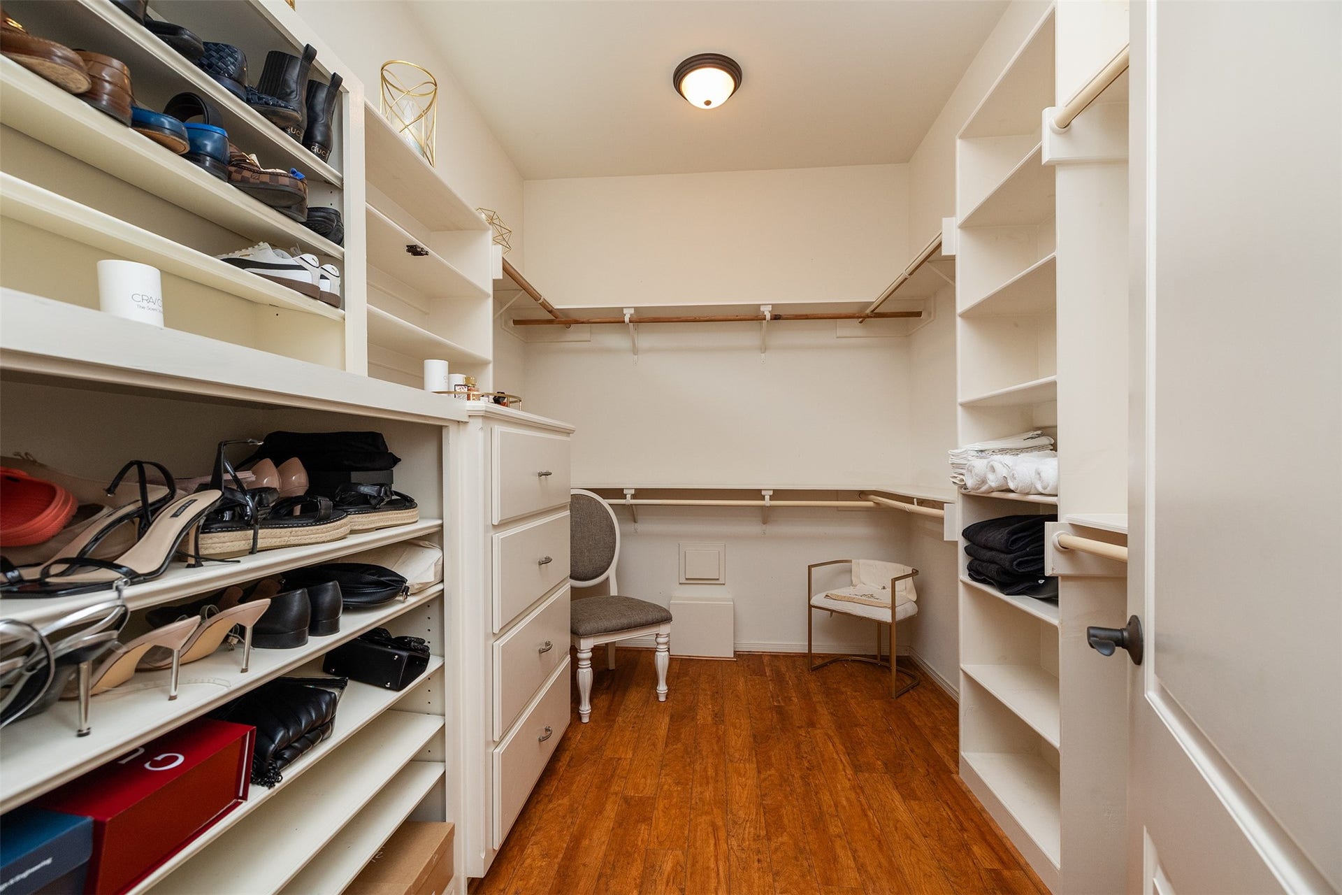 This photo showcases a spacious walk-in closet featuring ample shelving, drawers, and hanging space, with wooden flooring and a ceiling light fixture.