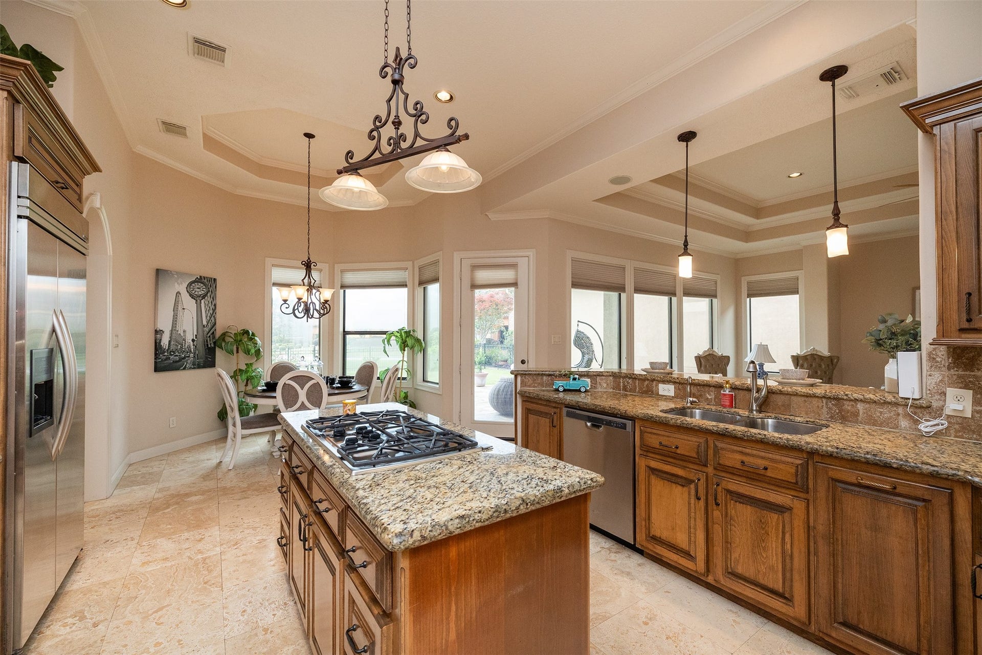 This spacious kitchen features elegant granite countertops, a central island with a gas cooktop, and rich wooden cabinetry. It opens to a bright dining area with large windows, offering plenty of natural light and a view of the outdoors.