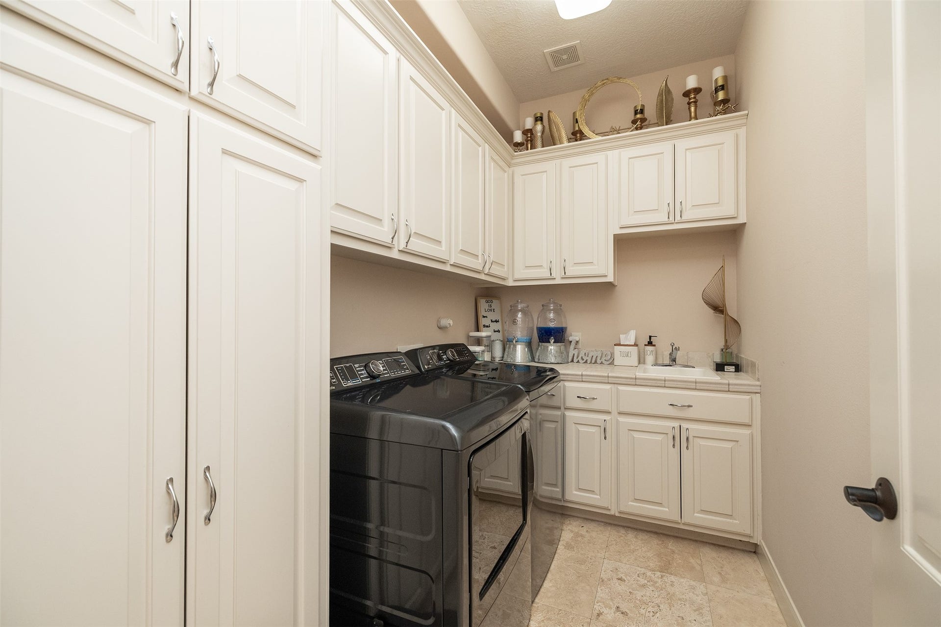 This photo showcases a well-organized laundry room featuring ample white cabinetry for storage, a modern black washer and dryer set, and a countertop for folding. The room is bright and functional, with decorative accents adding a touch of warmth.