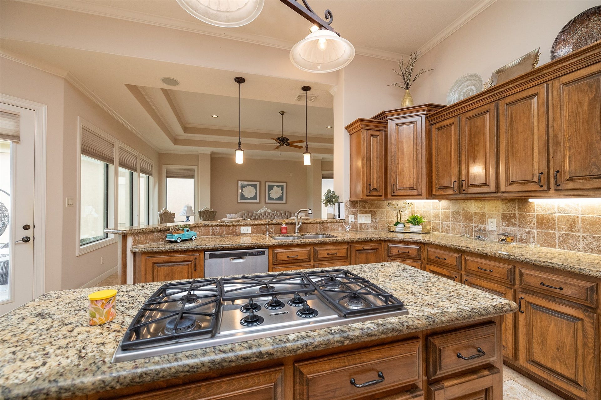 This kitchen features warm wood cabinetry, granite countertops, a center island with a gas cooktop, and ample natural light from large windows. The open layout includes a view into the adjacent living area, creating a spacious and inviting atmosphere.