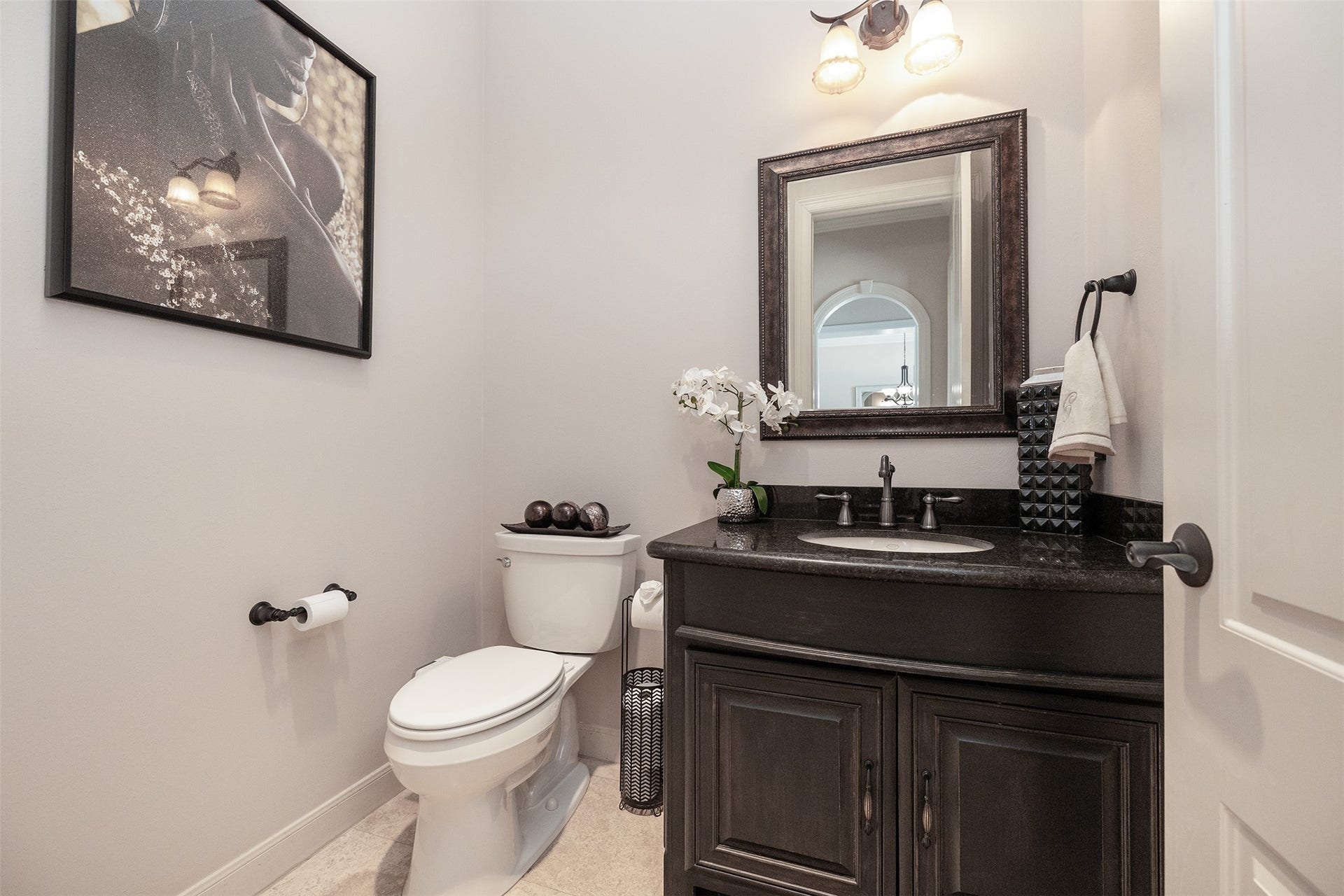 This photo showcases a stylish and modern half-bathroom with a dark wood vanity, sleek black countertop, elegant fixtures, and a decorative wall mirror. The light gray walls and tasteful artwork add a sophisticated touch.