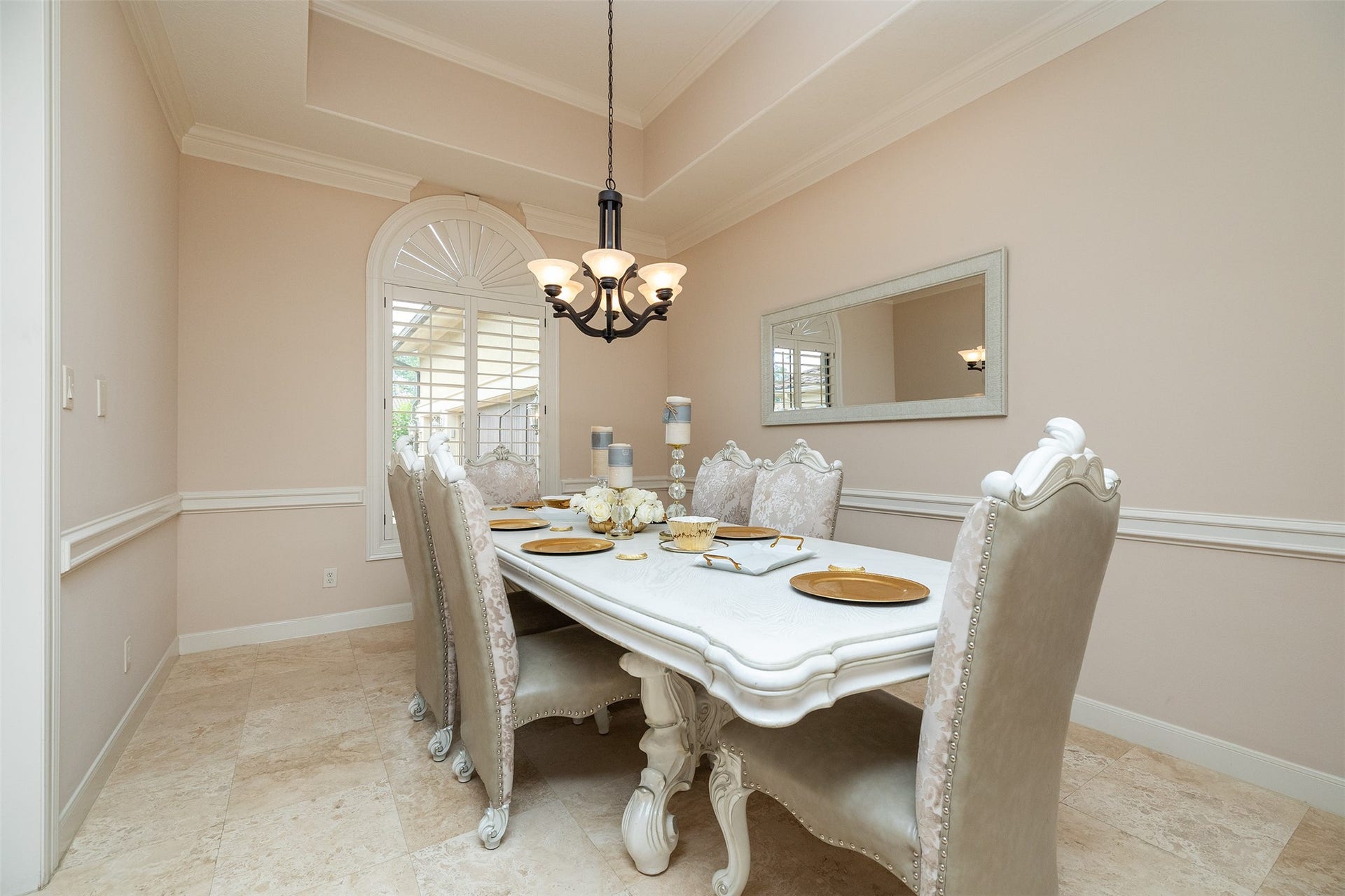 This elegant dining room features a classic chandelier, a large window with decorative shutters, and a stylish dining set with upholstered chairs. The neutral color palette and a wall mirror enhance the room's spacious feel.