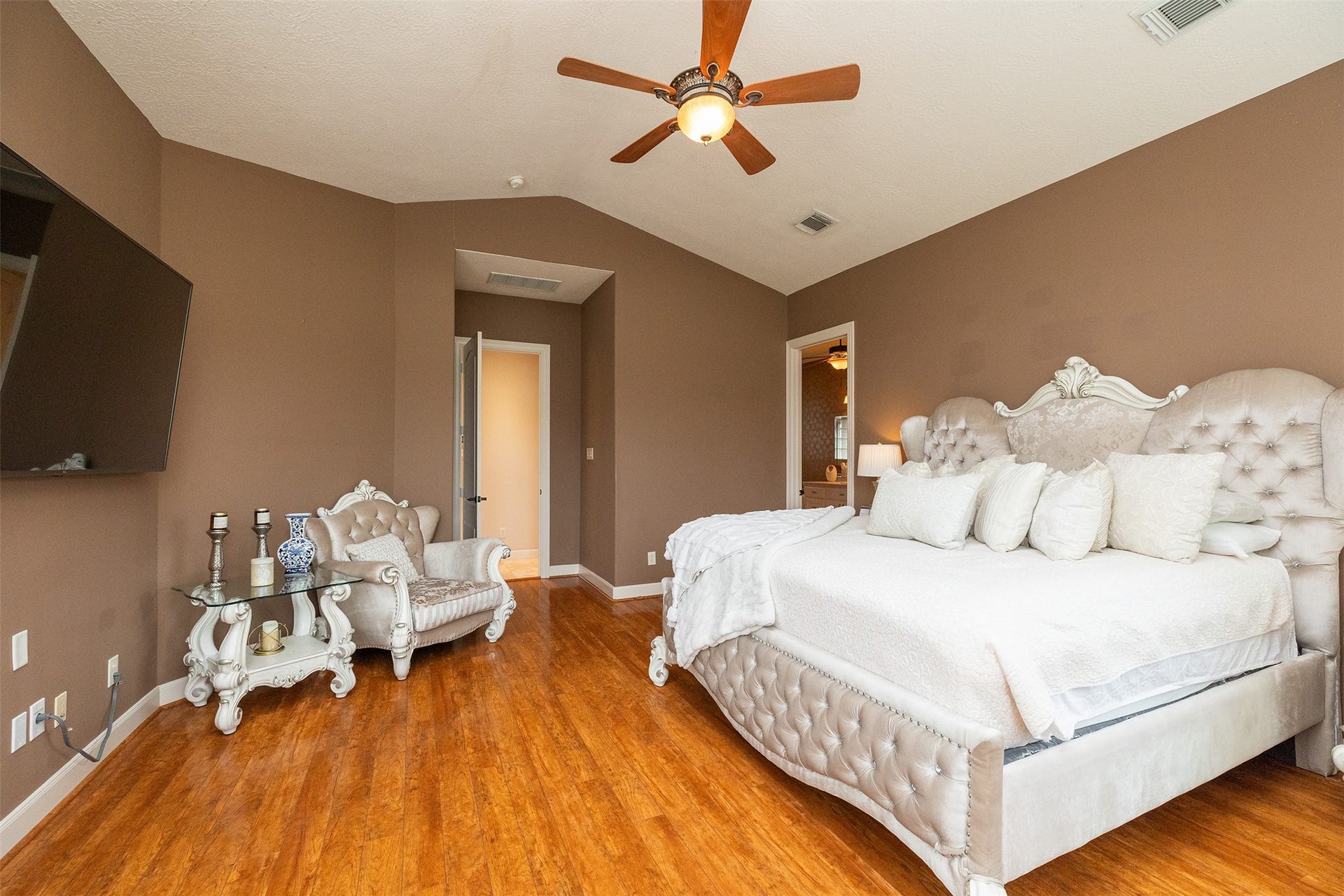 This spacious bedroom features warm brown walls, elegant wood flooring, and a high ceiling with a fan. It includes a luxurious tufted bed, a matching armchair, and a wall-mounted TV, creating a cozy and stylish retreat.