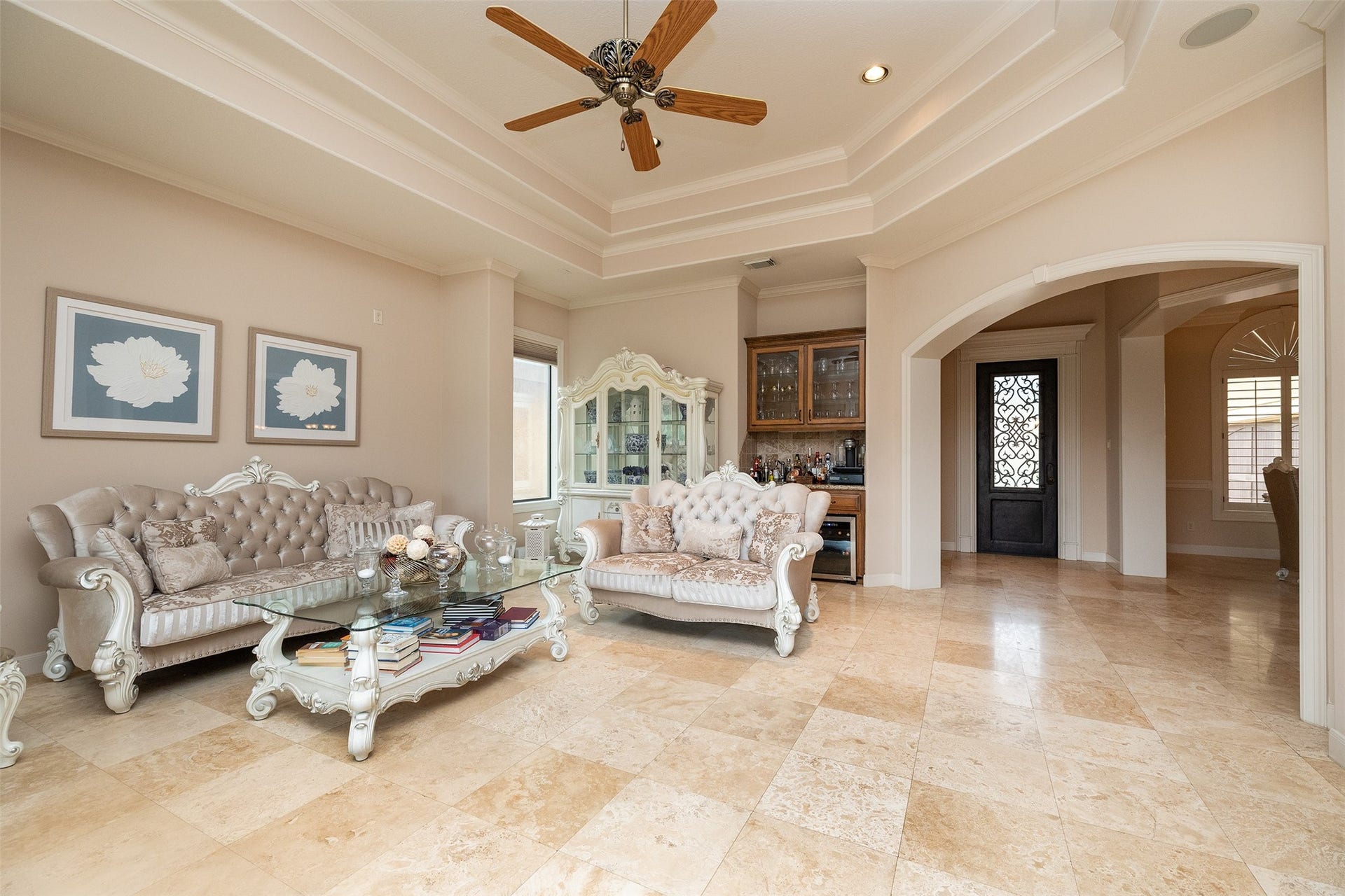 This elegant living room features high ceilings with crown molding, a ceiling fan, and polished tile floors. It includes ornate furniture, a glass coffee table, and decorative wall art. The space opens to a hallway with arched entryways and a stylish wrought iron door.