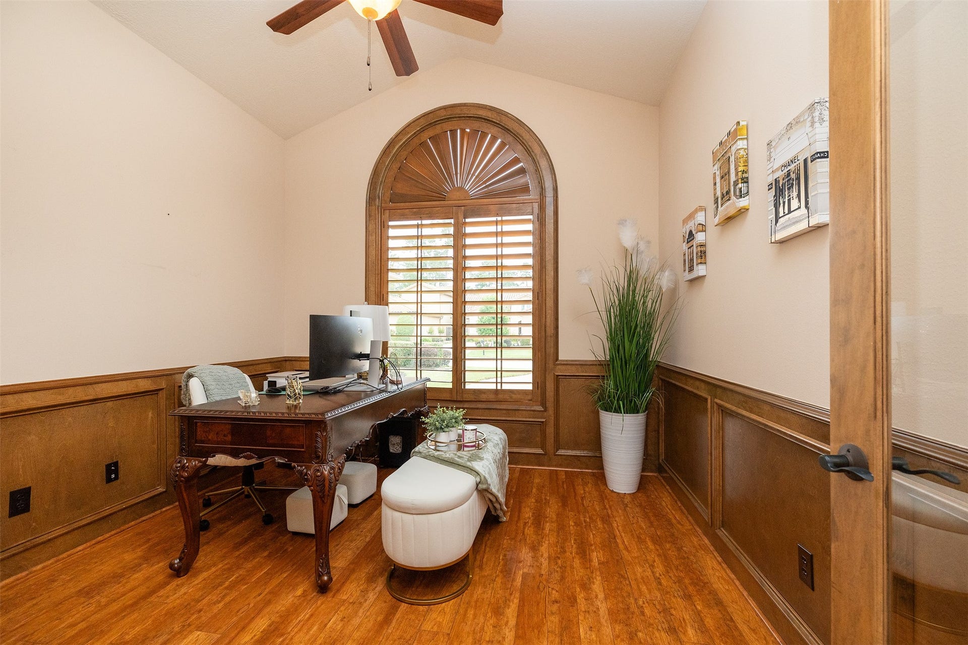 This room features a cozy home office with wood flooring, a large arched window with shutters, and elegant wainscoting. It includes a classic wooden desk, modern computer setup, and decorative plants, creating a warm and inviting workspace.