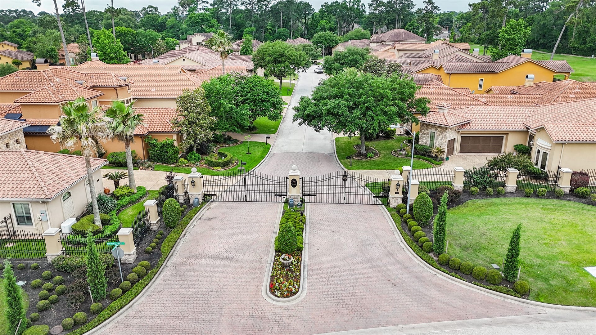 This photo showcases a gated community with beautifully landscaped gardens, Mediterranean-style homes, and tiled roofs. The entry is secure and elegant, surrounded by lush greenery and well-maintained pathways, offering a sense of privacy and exclusivity.