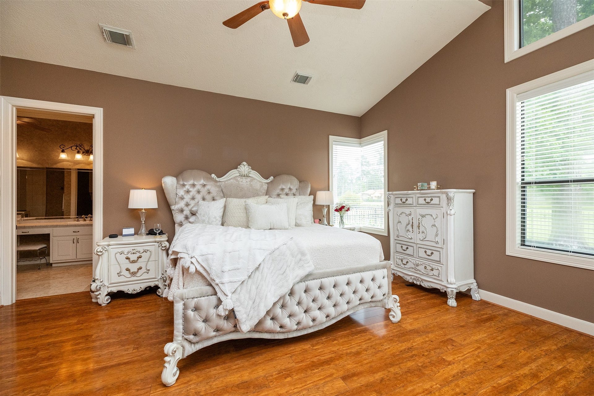 This spacious bedroom features elegant, tufted furniture with a vintage flair, warm brown walls, and rich wood flooring. Large windows provide ample natural light, and a ceiling fan enhances comfort. A doorway leads to an en-suite bathroom, adding convenience.