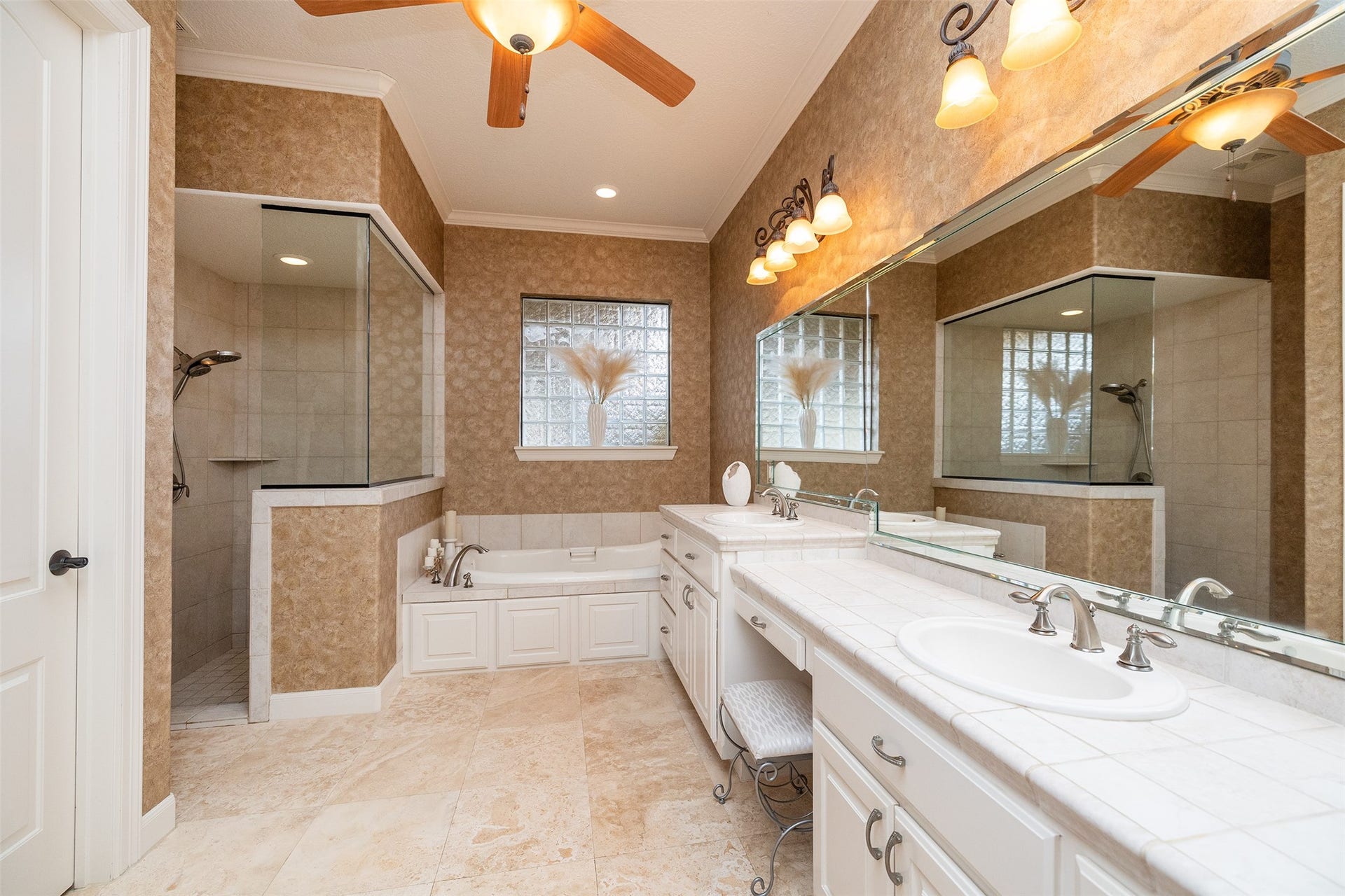 This bathroom features a spacious design with a walk-in shower, large bathtub, and dual sinks with elegant fixtures. The neutral tile flooring and warm lighting create a cozy atmosphere, complemented by a ceiling fan for added comfort.