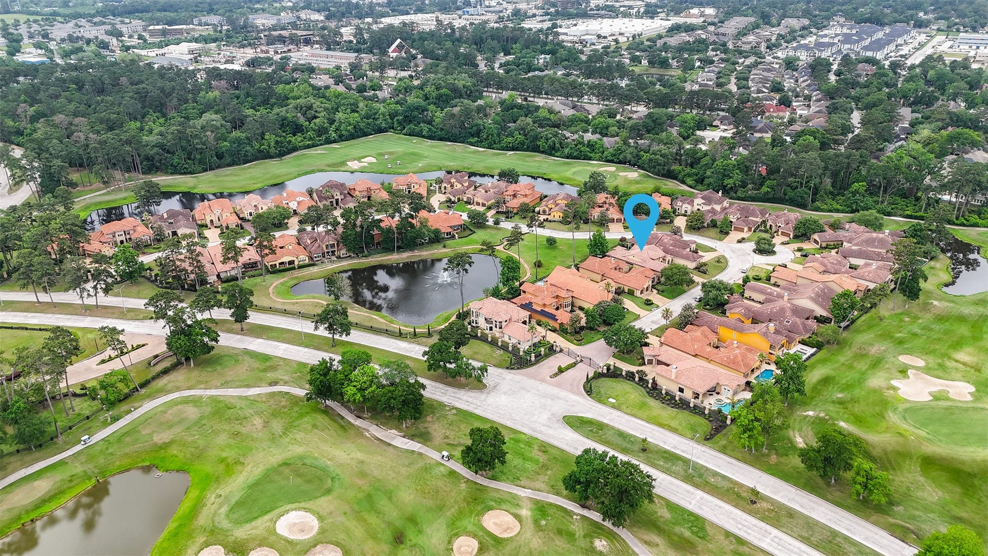 Aerial view of a serene residential community surrounded by lush greenery and a golf course. The neighborhood features elegant homes with terracotta roofs, water features, and is nestled near wooded areas, providing a peaceful and picturesque setting.