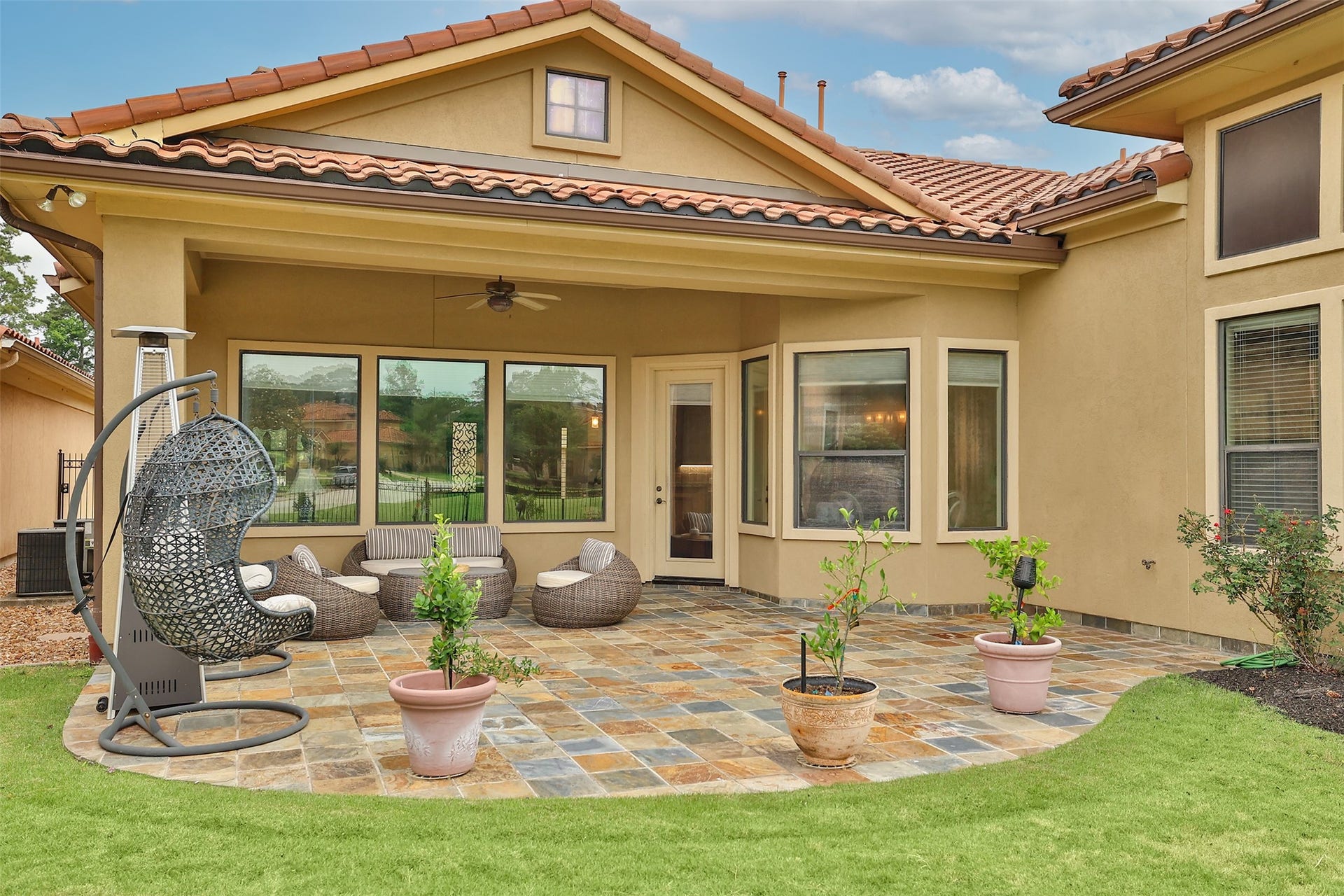This photo showcases a charming backyard patio with a tile floor, comfortable seating, a hanging chair, and potted plants. The area is covered, providing shade, and features large windows that offer views of the surrounding greenery. Ideal for relaxation and outdoor entertaining.