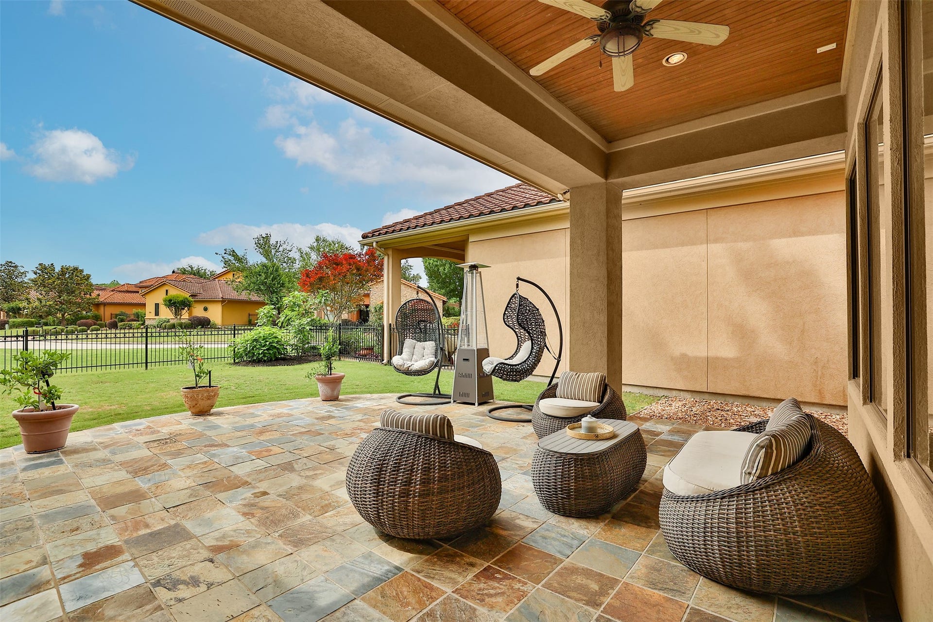 This photo showcases a spacious, tiled patio with stylish wicker furniture, a ceiling fan, and hanging chairs, perfect for outdoor relaxation. The area is surrounded by a well-maintained lawn and fenced yard, offering a peaceful view of neighboring homes.