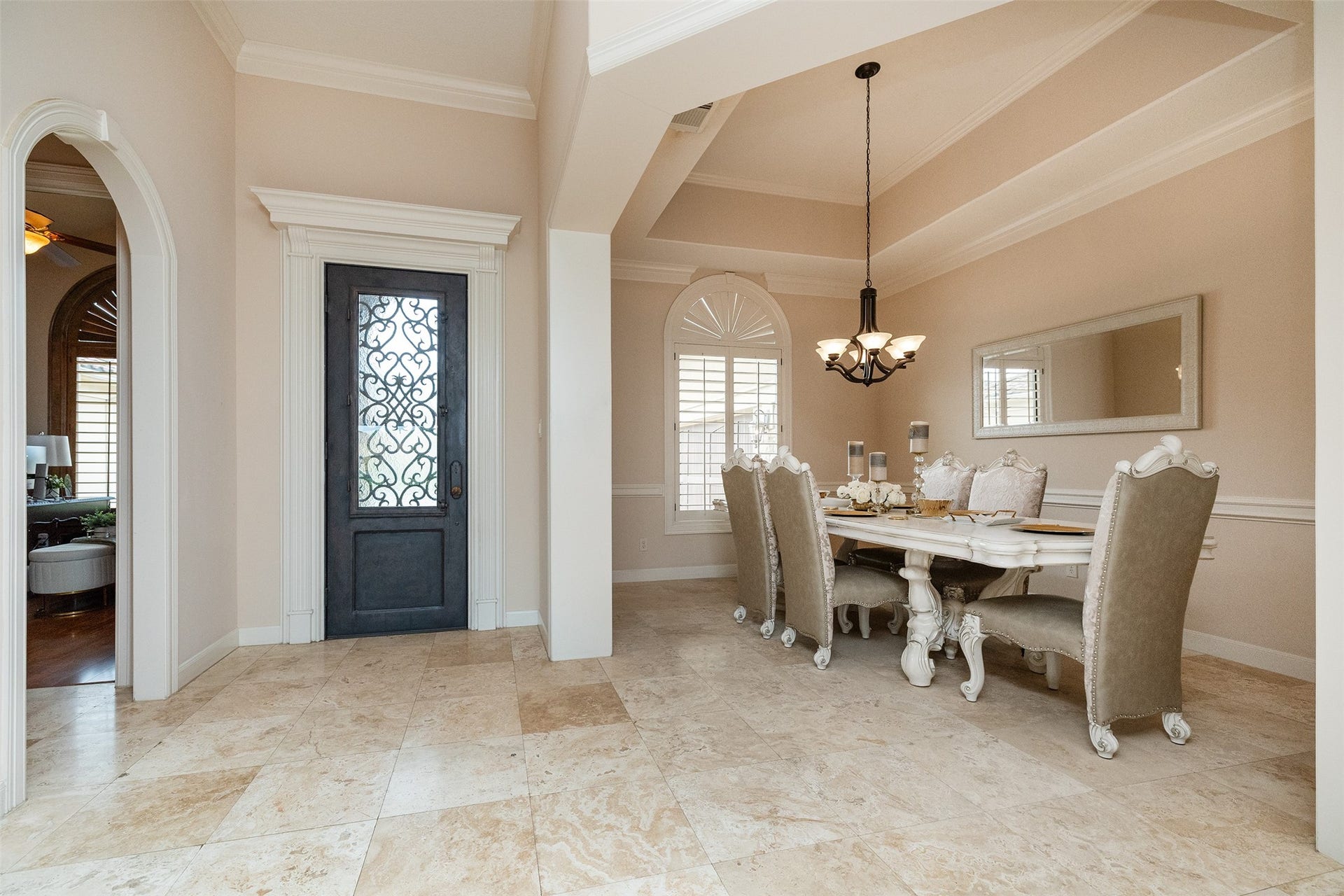 This elegant dining room features a coffered ceiling, a stylish chandelier, and a large window for natural light. The space is enhanced by a decorative wrought iron front door and neutral tile flooring, creating a sophisticated and inviting atmosphere.
