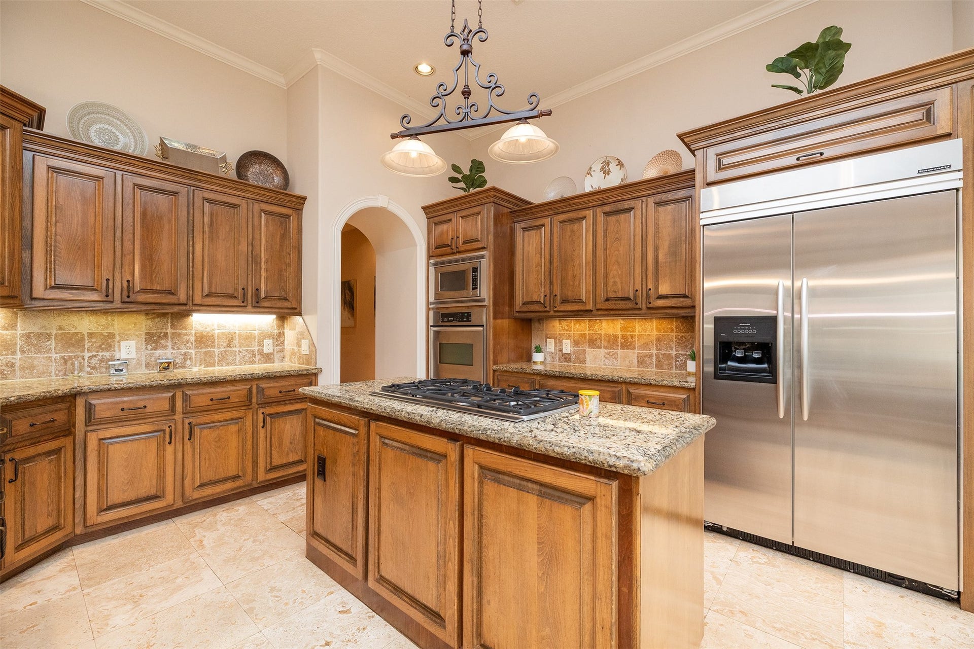 This kitchen features elegant wooden cabinetry with granite countertops, a central island with a built-in cooktop, stainless steel appliances, and tiled flooring. The space is accentuated by decorative lighting and ample storage.
