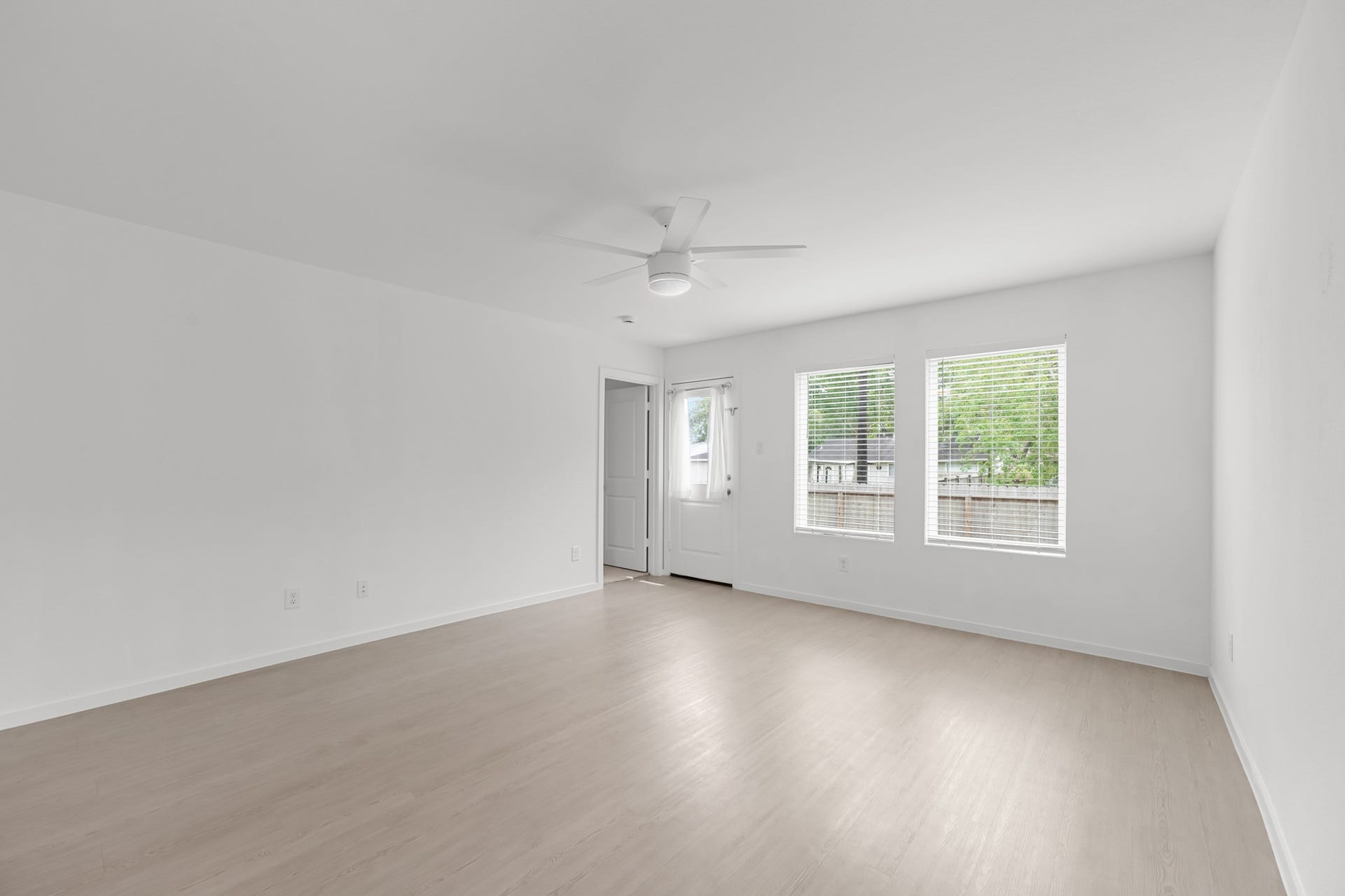 Bright living area features two windows, a ceiling fan, and light-toned flooring, with a rear door providing direct access to the outdoor space.