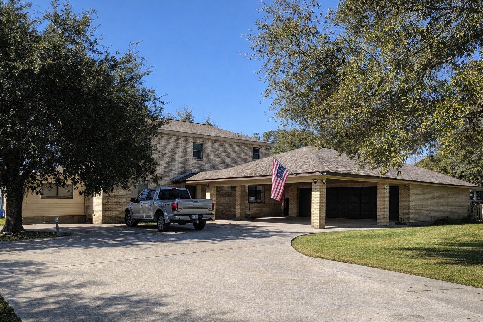 Impressive front elevation featuring brick exterior finishes, mature trees, and a spacious covered carport/porte-cochre parking area.