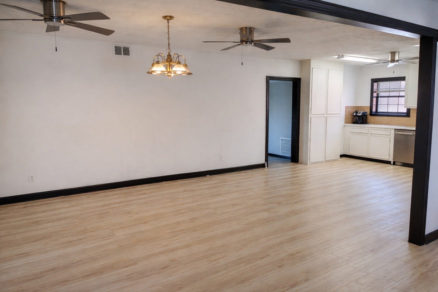 Expansive open living room featuring wood-look flooring, twin ceiling fans, classic chandelier lighting, and framed views into the adjacent kitchen.