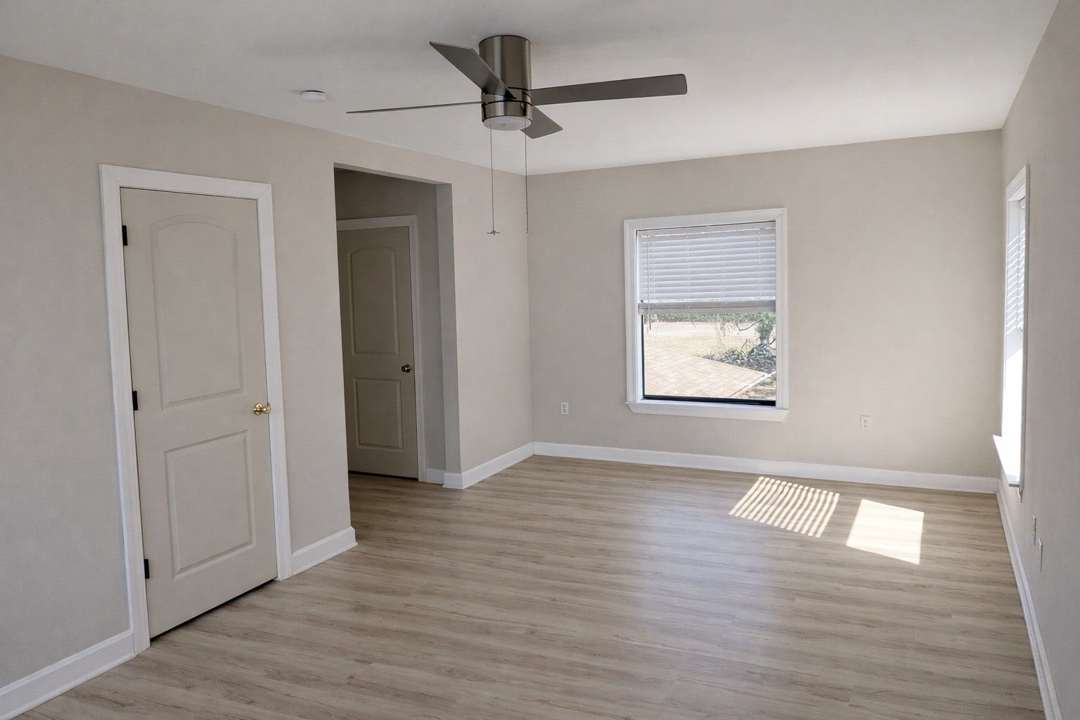 Well-proportioned bedroom offering light wood-look LVP flooring, neutral paint, a brushed-metal ceiling fan, and bright natural lighting.