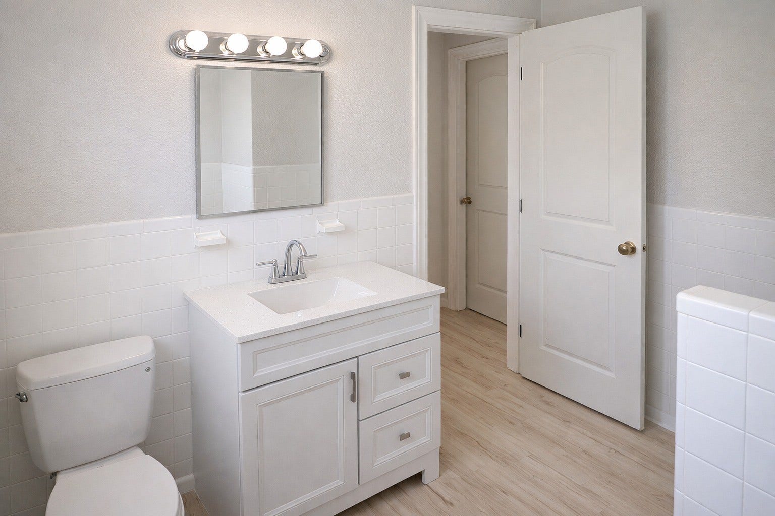 Bright updated bathroom featuring a white vanity with quartz countertop, vanity mirror lighting, light wood-look LVP flooring, and crisp white tile wainscoting.