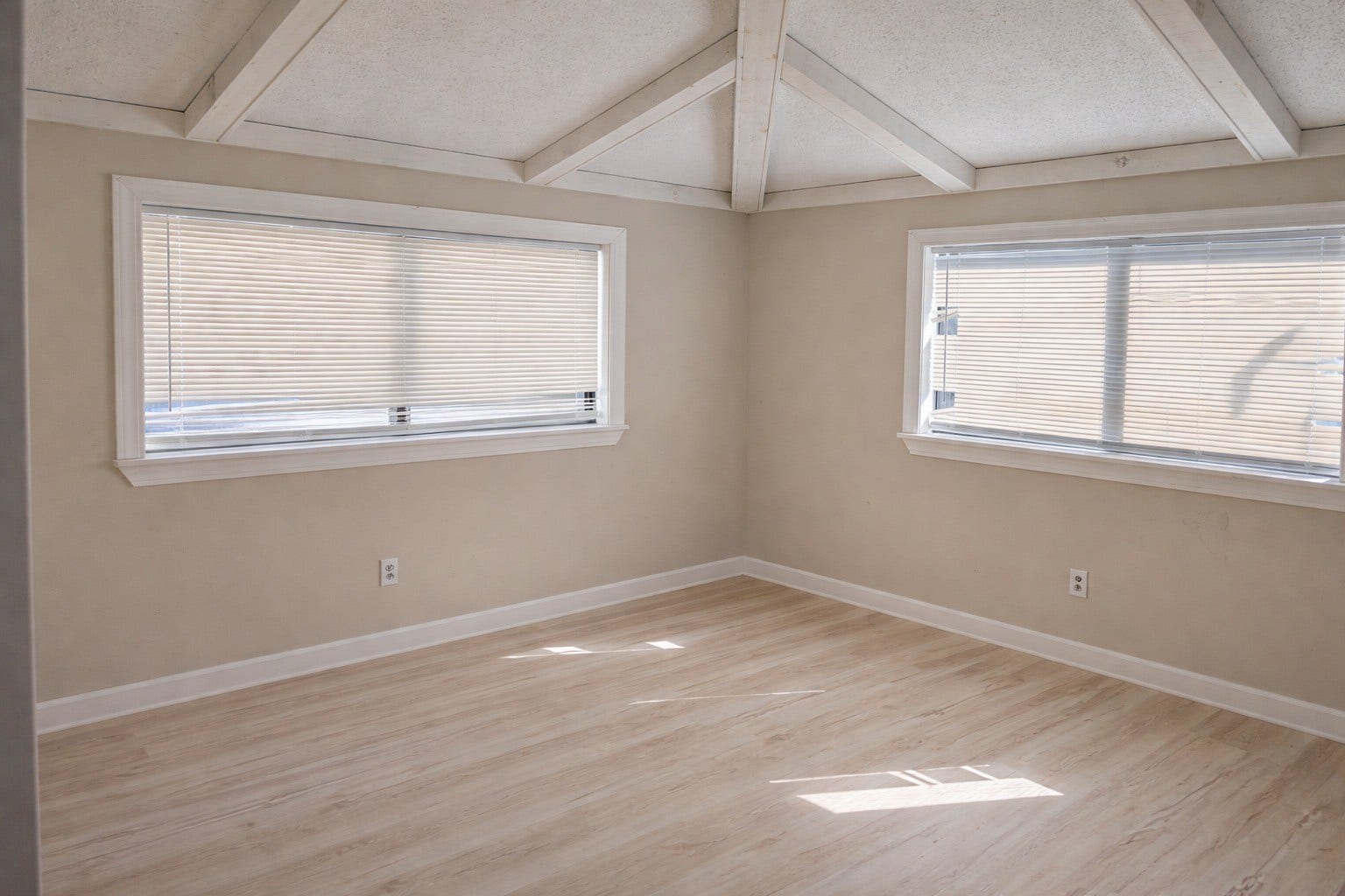 Well-lit bedroom room showcasing light wood-look flooring, double windows with horizontal blinds, and distinct vaulted ceiling beam details.