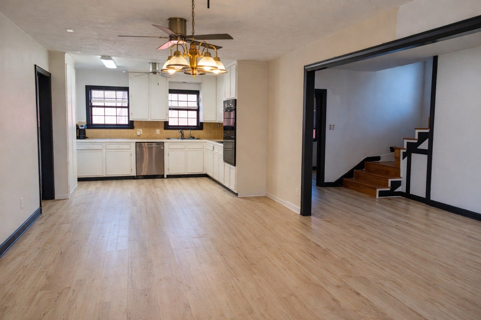 Open-concept living space with light-wood plank flooring, dark trim accents, a classic chandelier light fixture, and direct view into the functional kitchen layout.