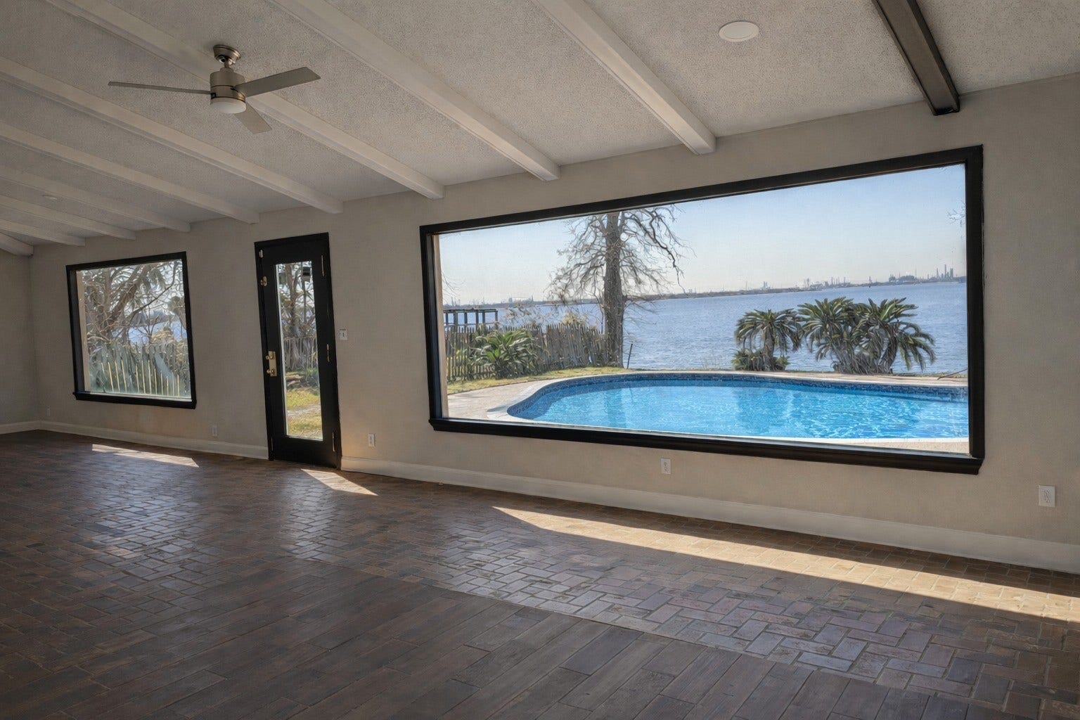 Expansive sunroom featuring brick-patterned tile flooring, exposed beam ceiling details, and sweeping picture windows framing pool and water views.