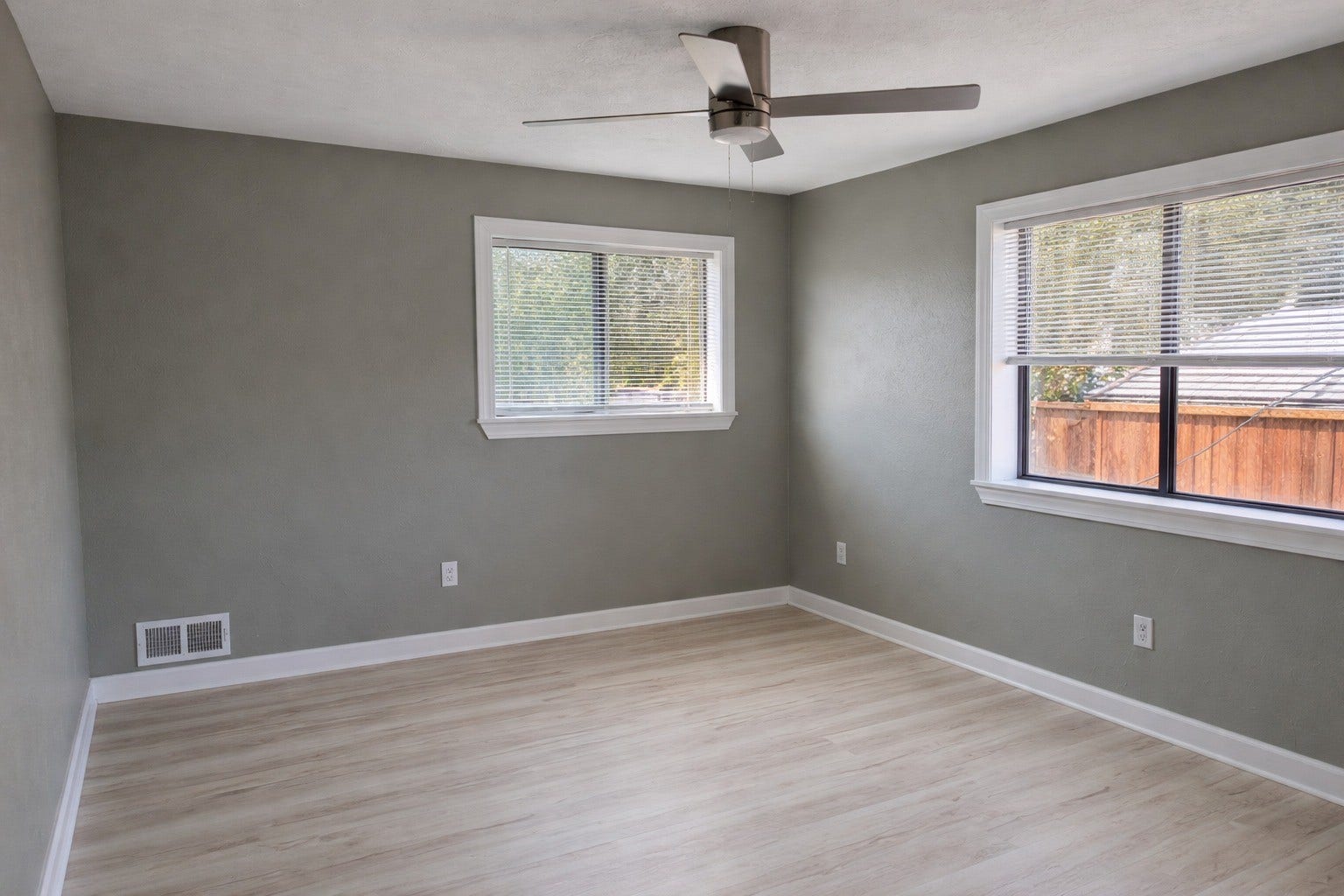 Spacious bedroom featuring crisp light-wood flooring, neutral gray walls, a modern brushed-metal ceiling fan, and large windows providing abundant natural light.