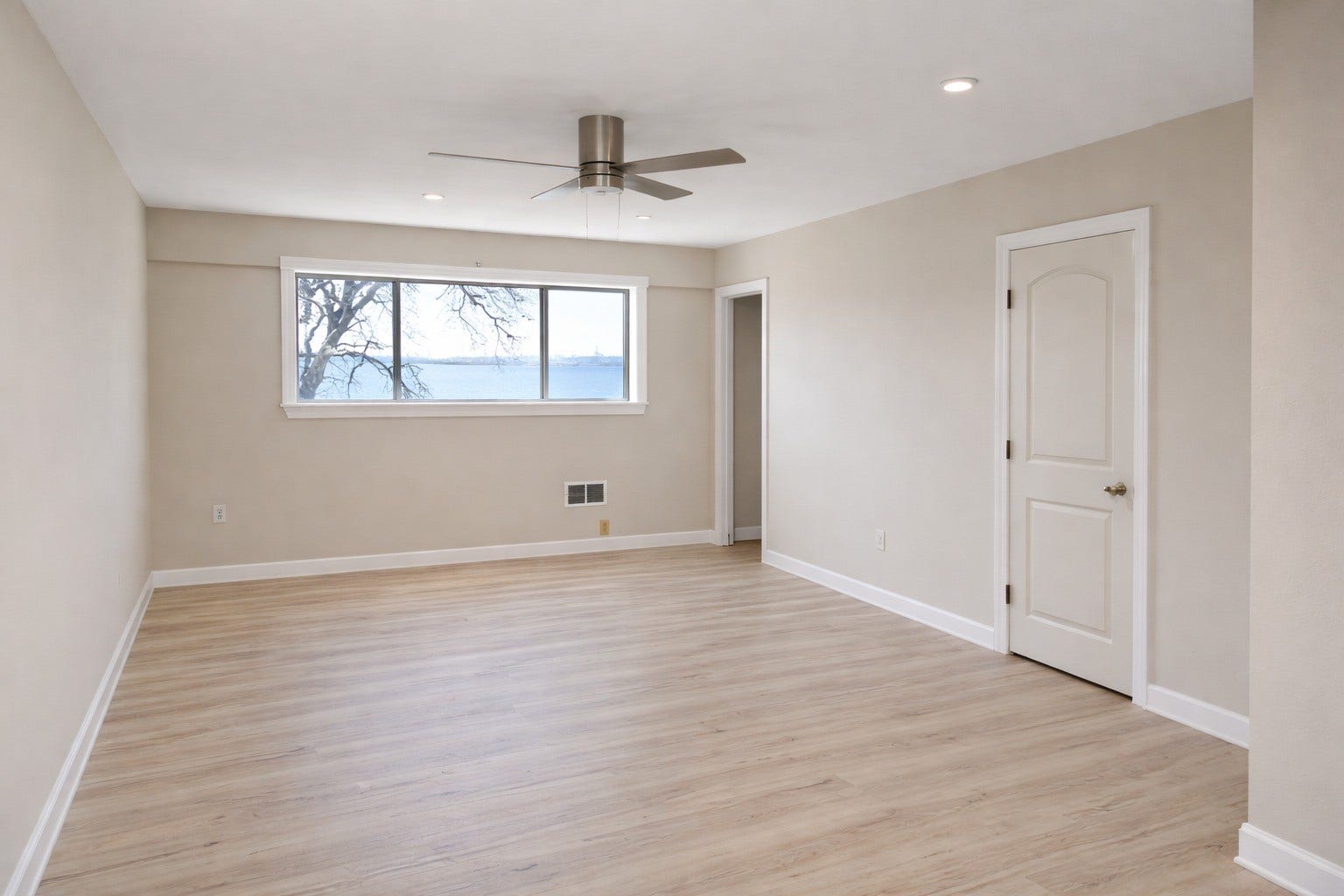 Expansive primary bedroom highlighting soft neutral wall tones, durable wood-look flooring, a sleek ceiling fan, and framed bay views.
