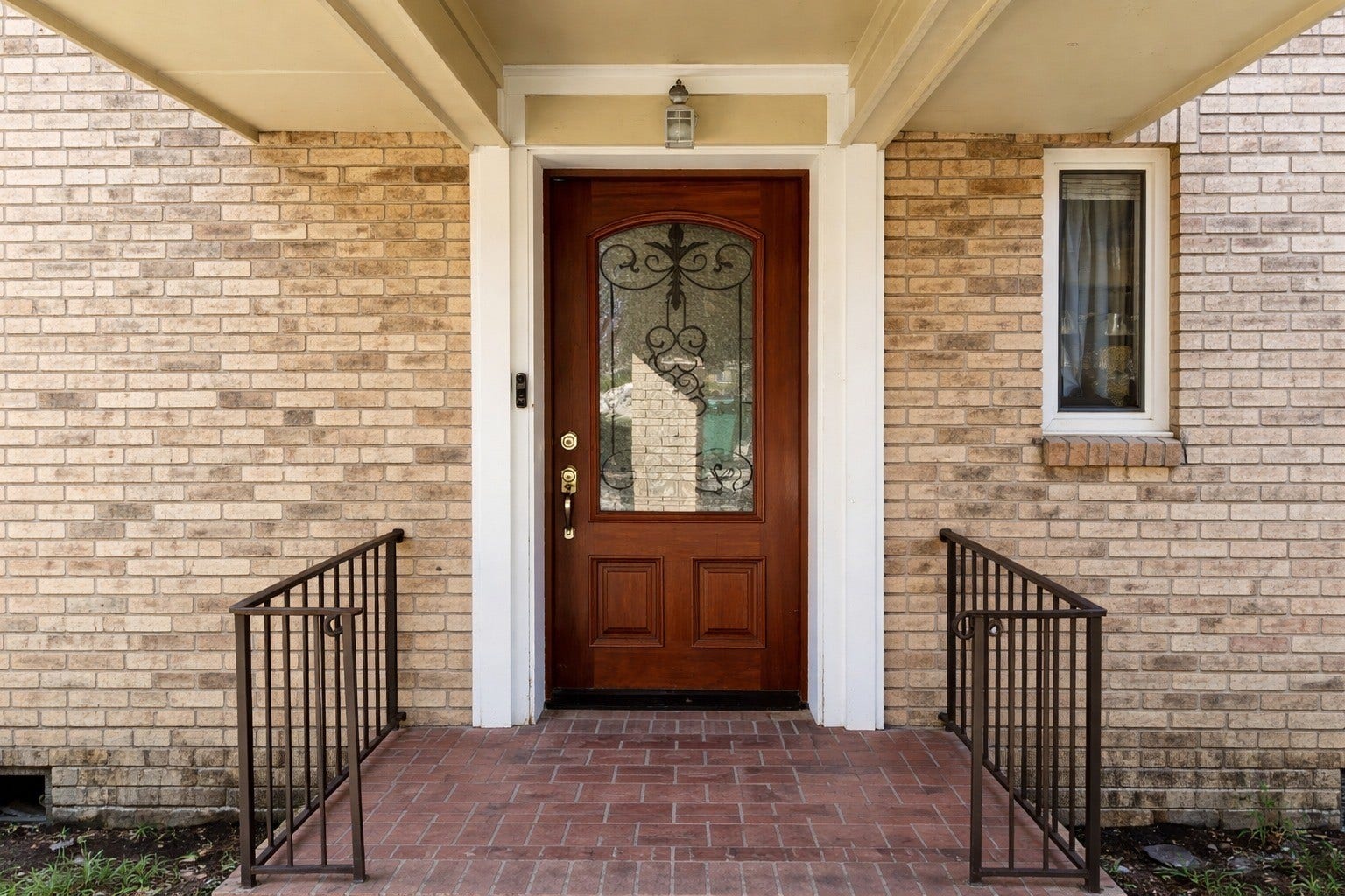 Covered front porch with brick paver flooring, wrought-iron railings, and an elegant wood front door with decorative glass inlay.