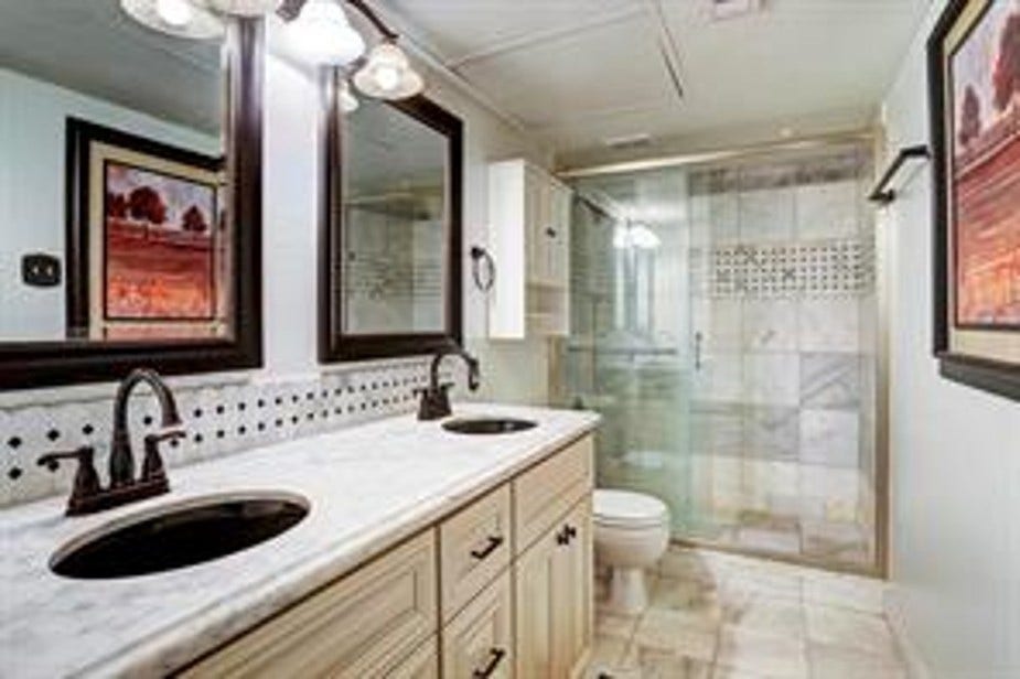 This bathroom features a dual vanity with elegant marble countertops and dark fixtures. The spacious walk-in shower has glass doors and stylish tile accents.