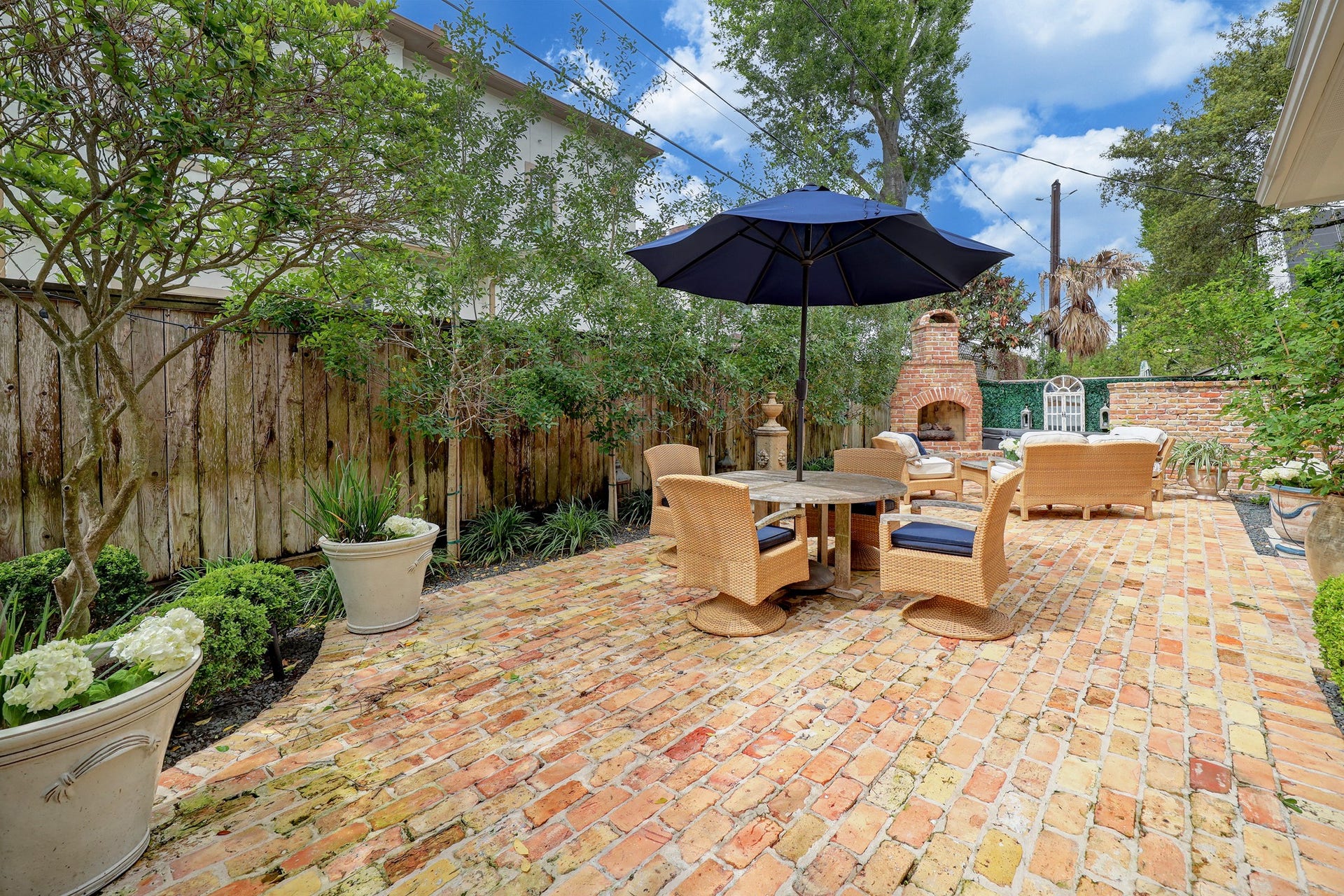 6004 Lake Street, Houston, TX 77005 Charming brick patio with cozy seating area, shaded by a large umbrella, features lush greenery and an outdoor fireplace for perfect entertaining.