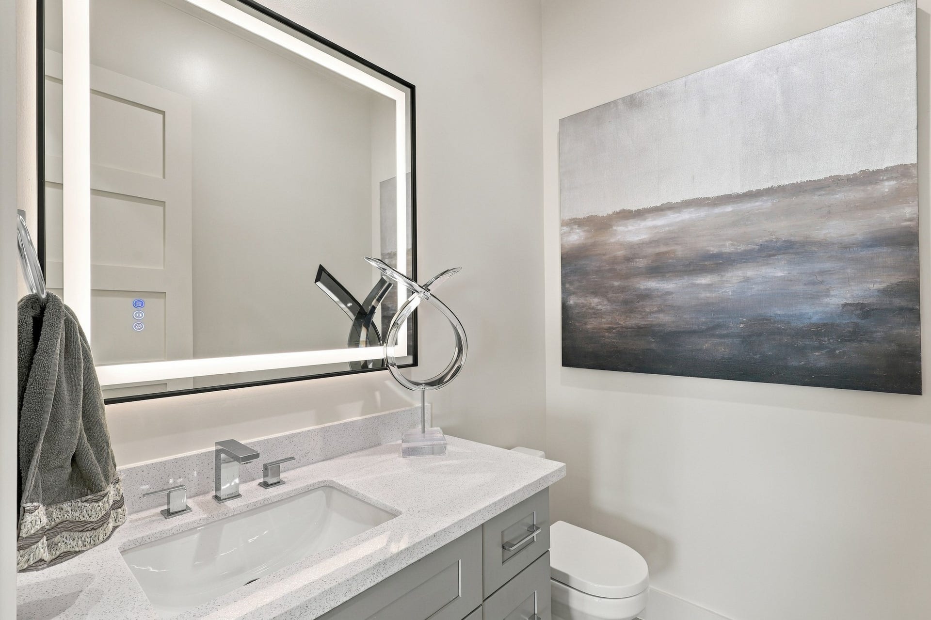 17211 Termini San Luis Pass Rd, Galveston, TX 77554 Stylish powder room/ Half Bath with a light countertop, gray vanity, contemporary chrome fixtures, and an illuminated mirror. Clean lines carry the homes modern aesthetic into this thoughtfully finished space.