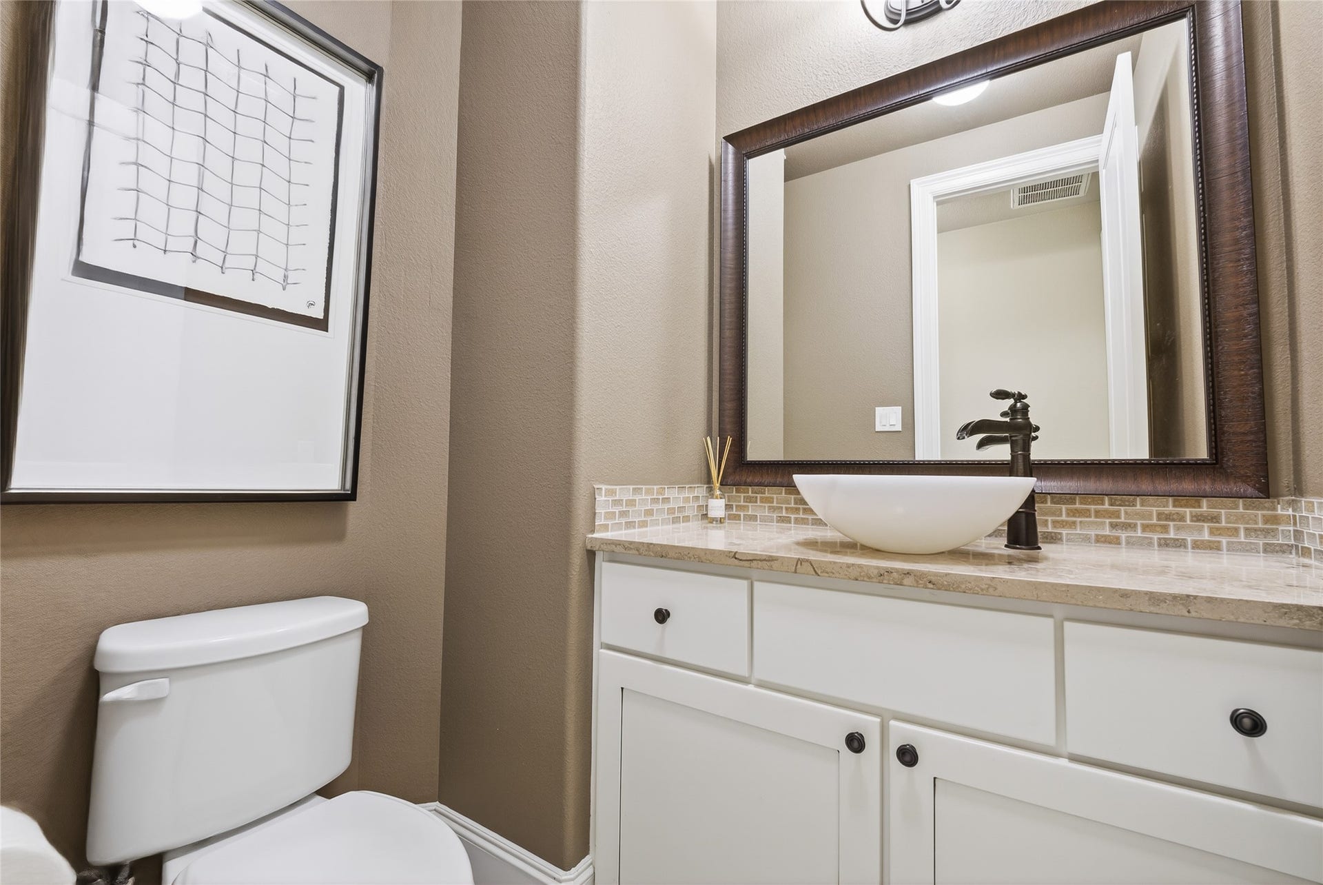 7907 Augusta Creek Court, Spring, TX 77389 This modern powder room features a sleek vessel sink, elegant faucet, and a large framed mirror. The neutral-toned walls, mosaic backsplash, and countertop create a warm, inviting atmosphere.