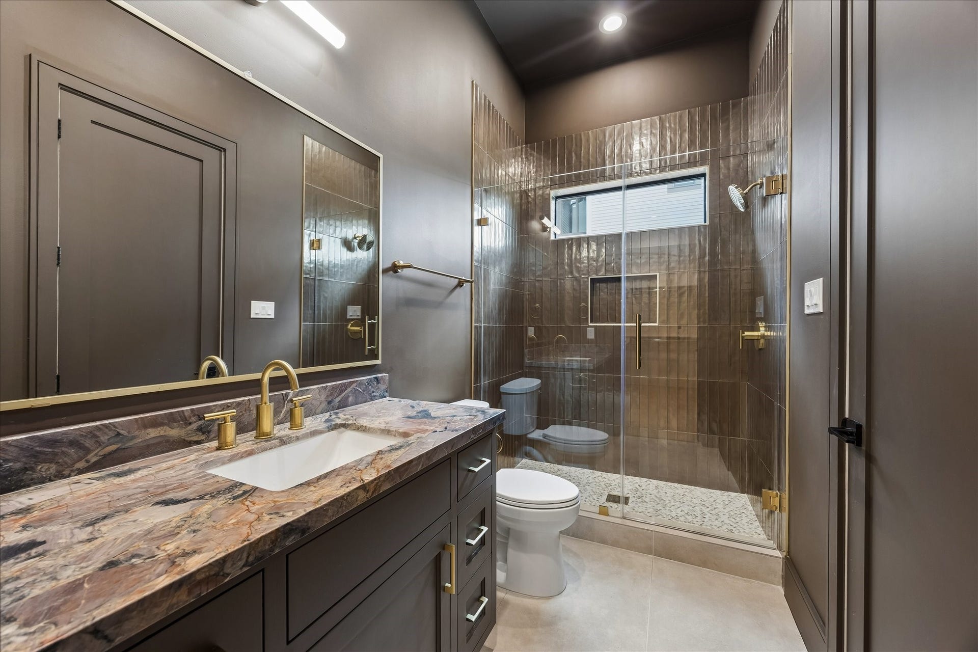316 W 30th Street, Houston, TX 77018 Rich stone surfaces, dramatic full height tile, and warm brass fixtures give this ensuite bath a distinctive designer feel. A frameless glass shower with built in niche and transom window completes the sophisticated space.