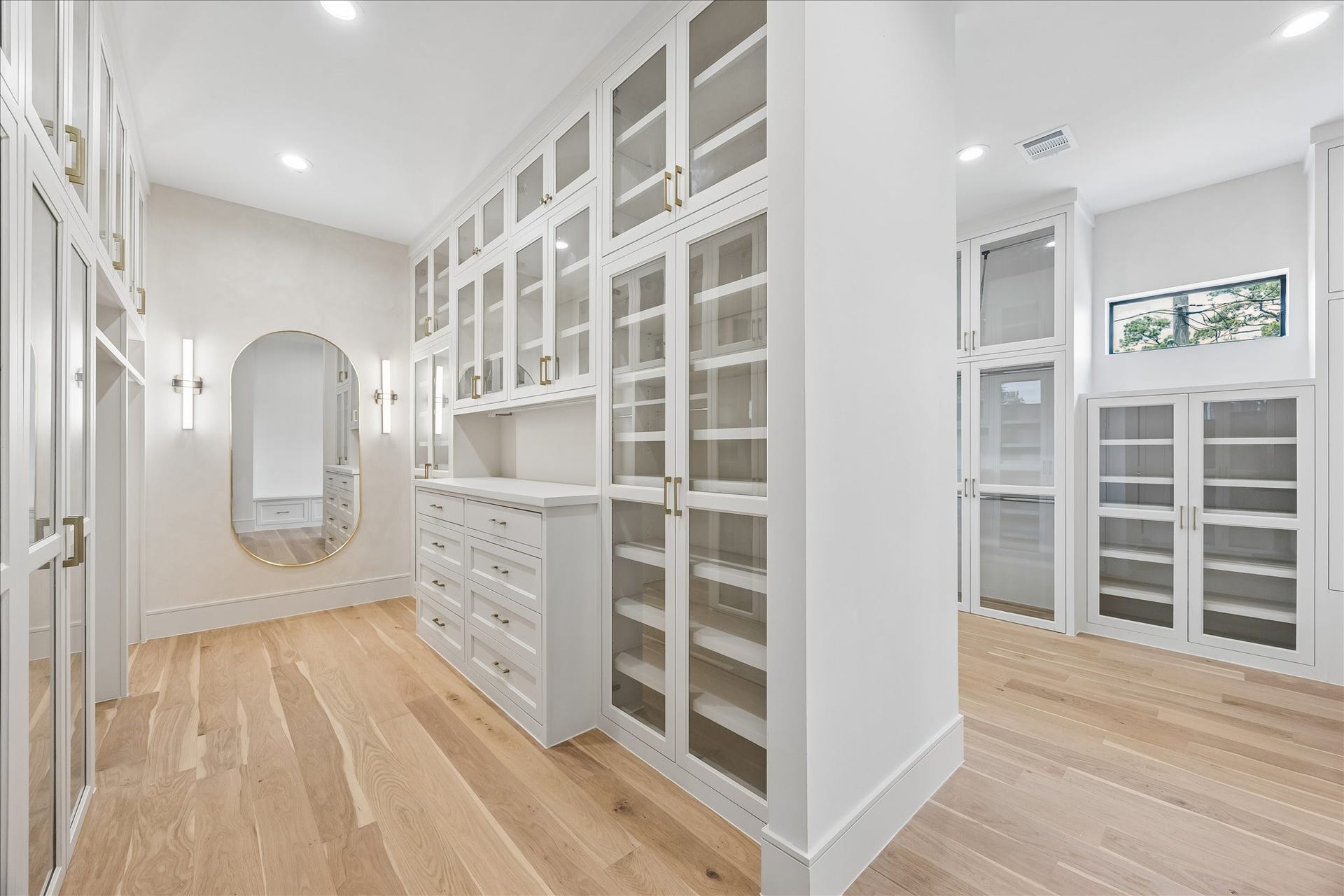 316 W 30th Street, Houston, TX 77018 Boutique style dressing room featuring floor to ceiling custom cabinetry, glass display doors, integrated drawer storage, and a center dressing station. The back wall is lime washed to match the homes soft white palette and finished with designer sconces and mirrors, with all cabinetry pre-wired for LED lighting to be added as an upgrade. Thoughtfully designed with abundant storage, natural light, and direct access to the spa inspired primary bath.