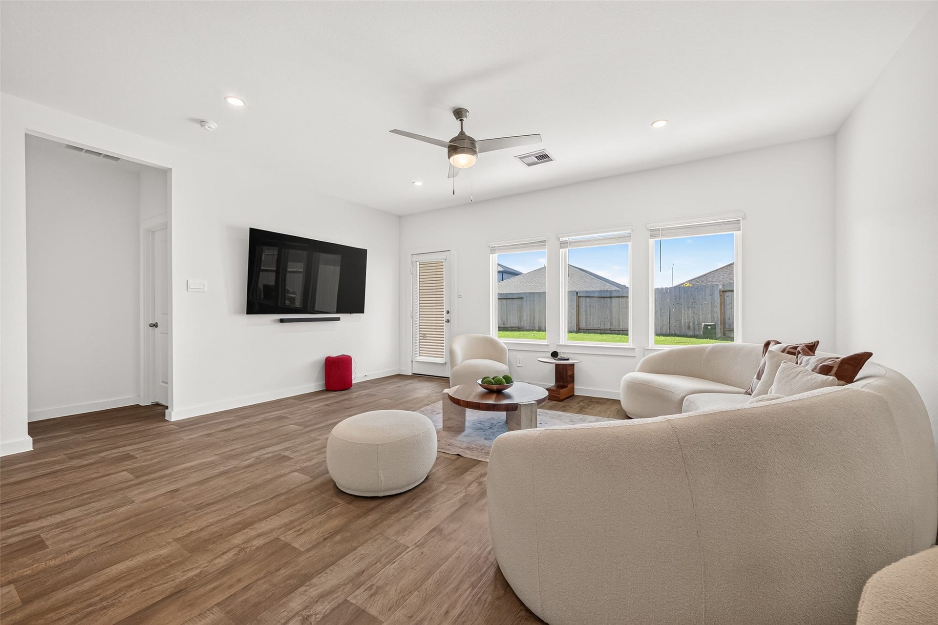Bright and inviting living room with sleek wood flooring, modern ceiling fan, and ample natural light from large windows. Perfect for relaxation with its cozy seating and wall-mounted TV.
