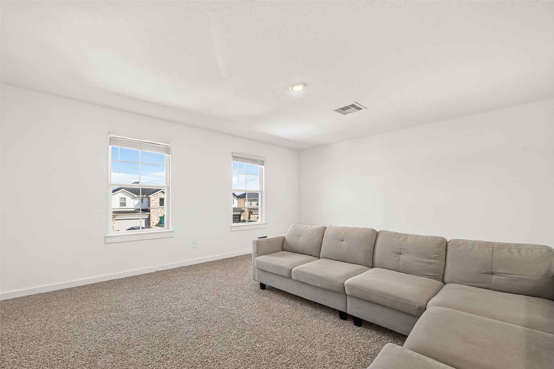 Bright and spacious living area with plush carpeting, abundant natural light from two large windows, and a cozy sectional sofa perfect for relaxation.