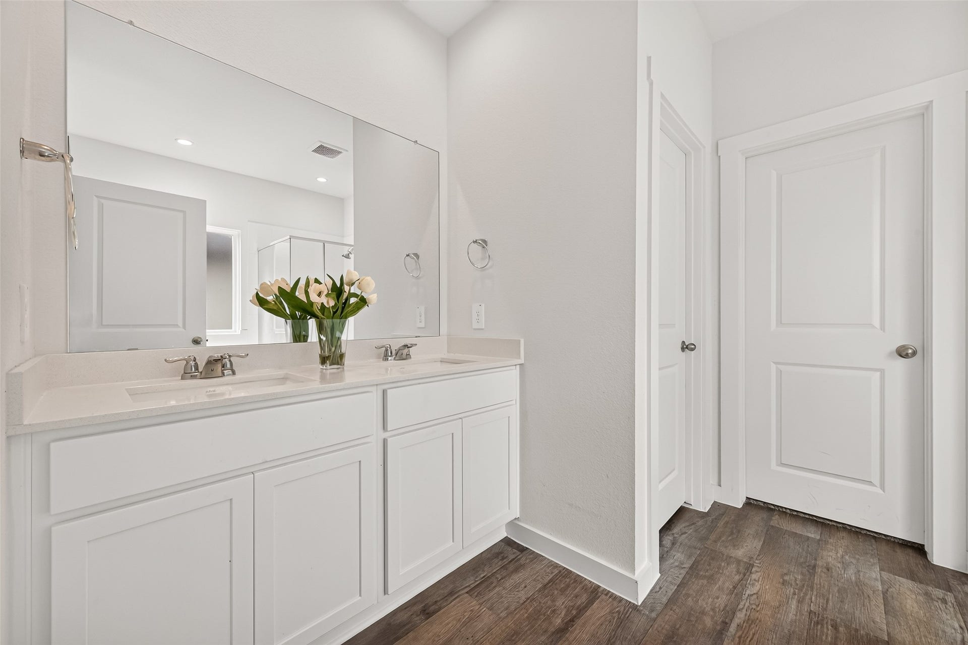 Elegant bathroom featuring a dual-sink vanity with modern fixtures, complemented by sleek white cabinetry and a large mirror, all set on stylish wood-look flooring.